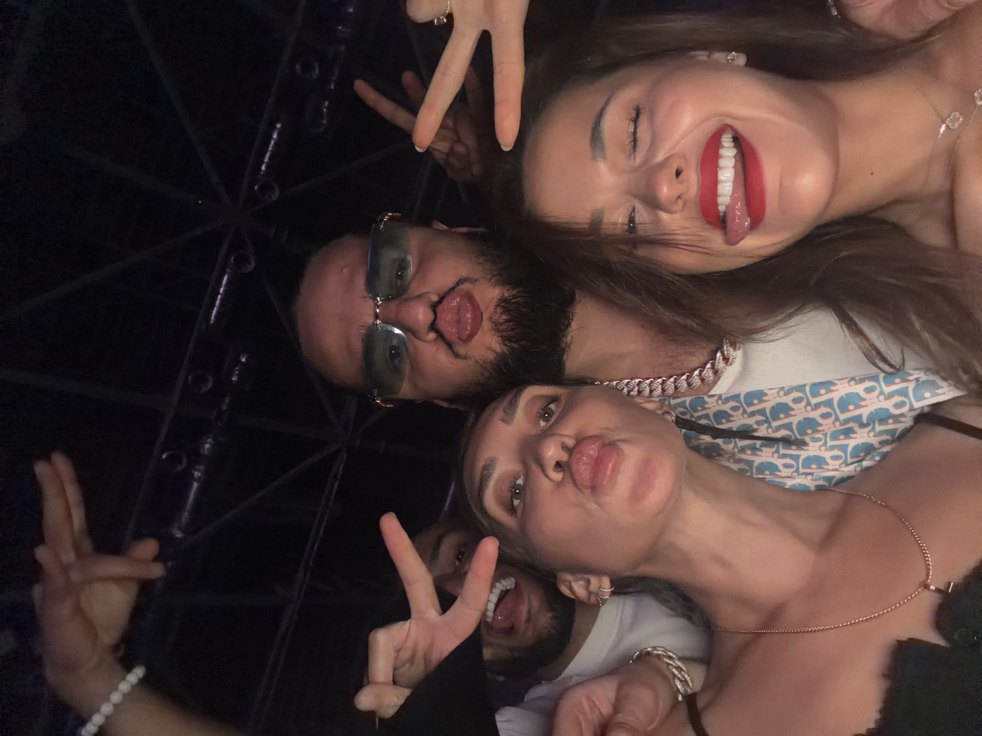 This image, reportedly taken in Khimki, Russia, captures a close-up group selfie featuring at least four individuals in a dimly lit indoor environment, characteristic of a nightclub or social event. The background reveals dark structural elements, possibly overhead trusses or lighting fixtures.
In the foreground, two women are prominent. The woman on the left, with dark hair, wears a dark, textured top and a gold chain necklace with an initial 'T' pendant. She poses with pursed lips, forming a "kissing" face. The woman on the right, also with dark hair, has red lipstick, a wide smile with her tongue playfully extended, and her eyes squinted. She wears a gold necklace with two clover-shaped pendants and a stud earring.
Positioned centrally behind them is a man with a beard and dark hair, wearing large rectangular blue-tinted sunglasses with gold frames and a substantial silver-colored Cuban link chain. He makes a "fish face" with pursed lips and a slightly extended tongue. To the left, another bearded man is partially visible, laughing with an open mouth.
Multiple hands are visible within the frame, making "peace signs" (V-gestures) around the group. One hand in the upper-left wears a white beaded bracelet, and a hand in the lower-left wears a silver-colored chain bracelet. A patterned blue and white scarf with a repeated monogram (resembling 'CD') is draped over the central man's shoulder, next to the woman on the left. The group appears engaged in playful interaction for the camera.