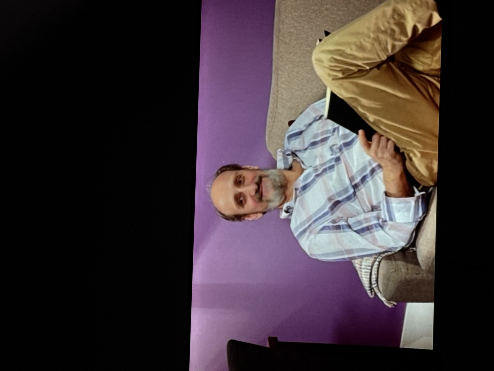 A middle-aged man with a bald head and beard is sitting on a couch, holding a book and smiling. He is wearing a plaid shirt and khaki pants. The couch is light brown and appears to be in a living room with a purple wall in the background. There is a black chair to the left of the man. The image is a photograph taken from a slight angle, with the top and bottom portions of the image appearing blacked out. There is no information to suggest this image is located in Delmenhorst, Germany.