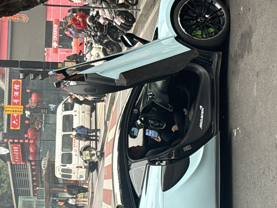 A light blue McLaren supercar with a raised door sits on the street. Inside, a person in dark clothing is looking at a smartphone. The car is parked on a paved surface. In the background are people, parked motorcycles, a white van, and buildings with Chinese signage. Lanterns hang from a building in the background. The scene appears to be a bustling city street.