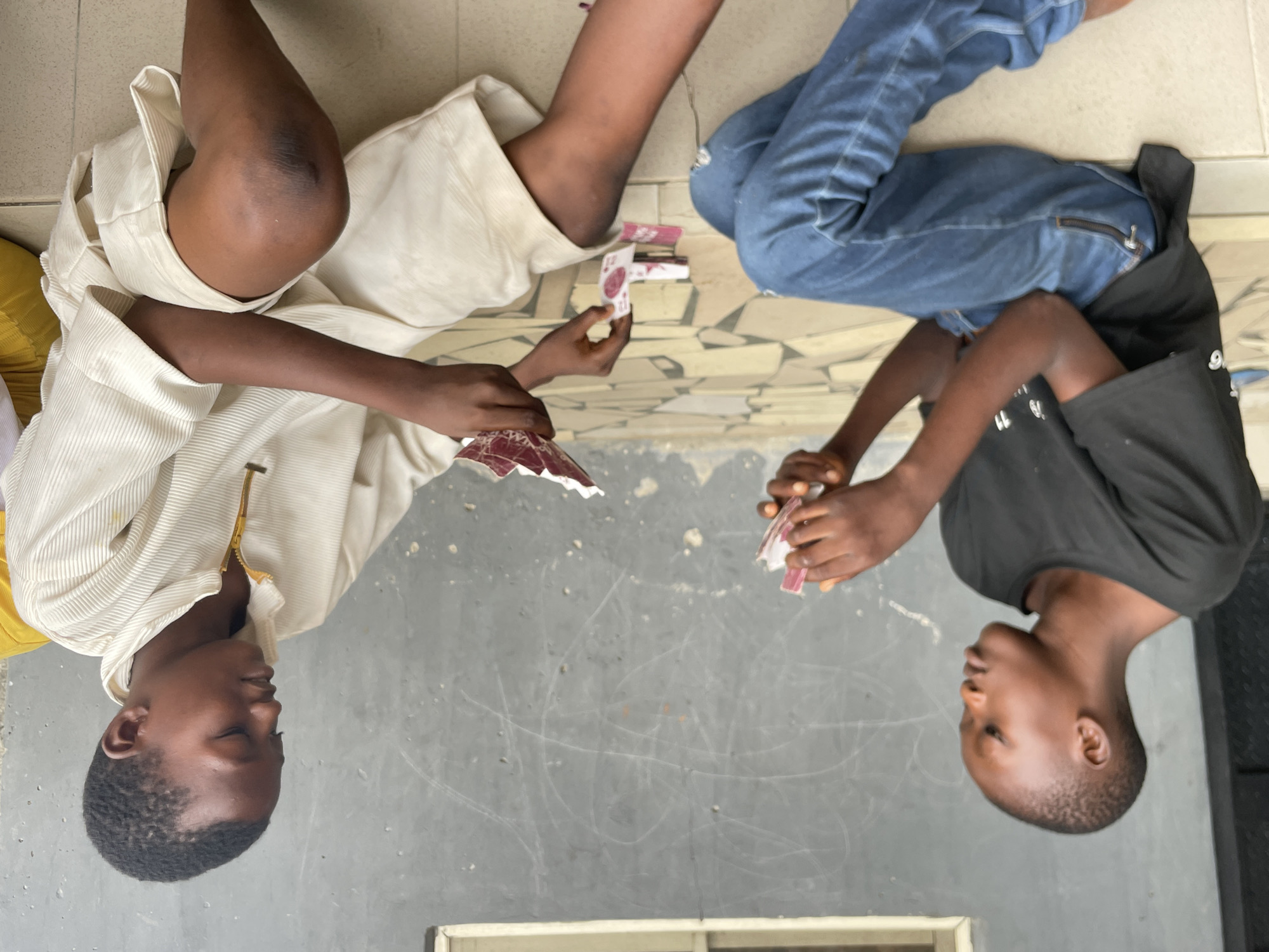 The image captures two young individuals, likely children or pre-teens, lying on the ground and engaged in a game involving small, rectangular, card-like objects. The scene is viewed from an overhead perspective, with the subjects facing each other.
On the left, one individual with dark skin and short dark hair is dressed in a light-colored, possibly cream, long-sleeved shirt or jacket featuring a gold-colored zipper, worn over a yellow undergarment, and light-colored pants. This person holds several small, patterned cards, which are predominantly red/maroon and white, in their left hand, seemingly examining them or their hand for the game. Their limbs are casually bent as they recline.
Opposite them, on the right, another individual with dark skin and short dark hair is wearing a black short-sleeved t-shirt and blue jeans, which appear to have distressed or ripped knees. This person holds a stack of similar small, patterned cards in their clasped hands. A silver-colored zipper is visible on their jeans. A small pile of these same cards rests on the ground between the two players.
The setting appears to be a casual outdoor or semi-outdoor space. The ground beneath the individual on the left consists of light-colored geometric tiles, transitioning to a darker grey concrete or paved surface with white scuff marks or drawings beneath the individual on the right and in the central area. A partial view of a window frame or similar structure is visible at the very bottom. The lighting suggests it is daytime, and the individuals appear calm and focused on their game, exhibiting no strong emotional expressions. The activity takes place in Badagry, Nigeria. No discernible text is visible on the cards or clothing.