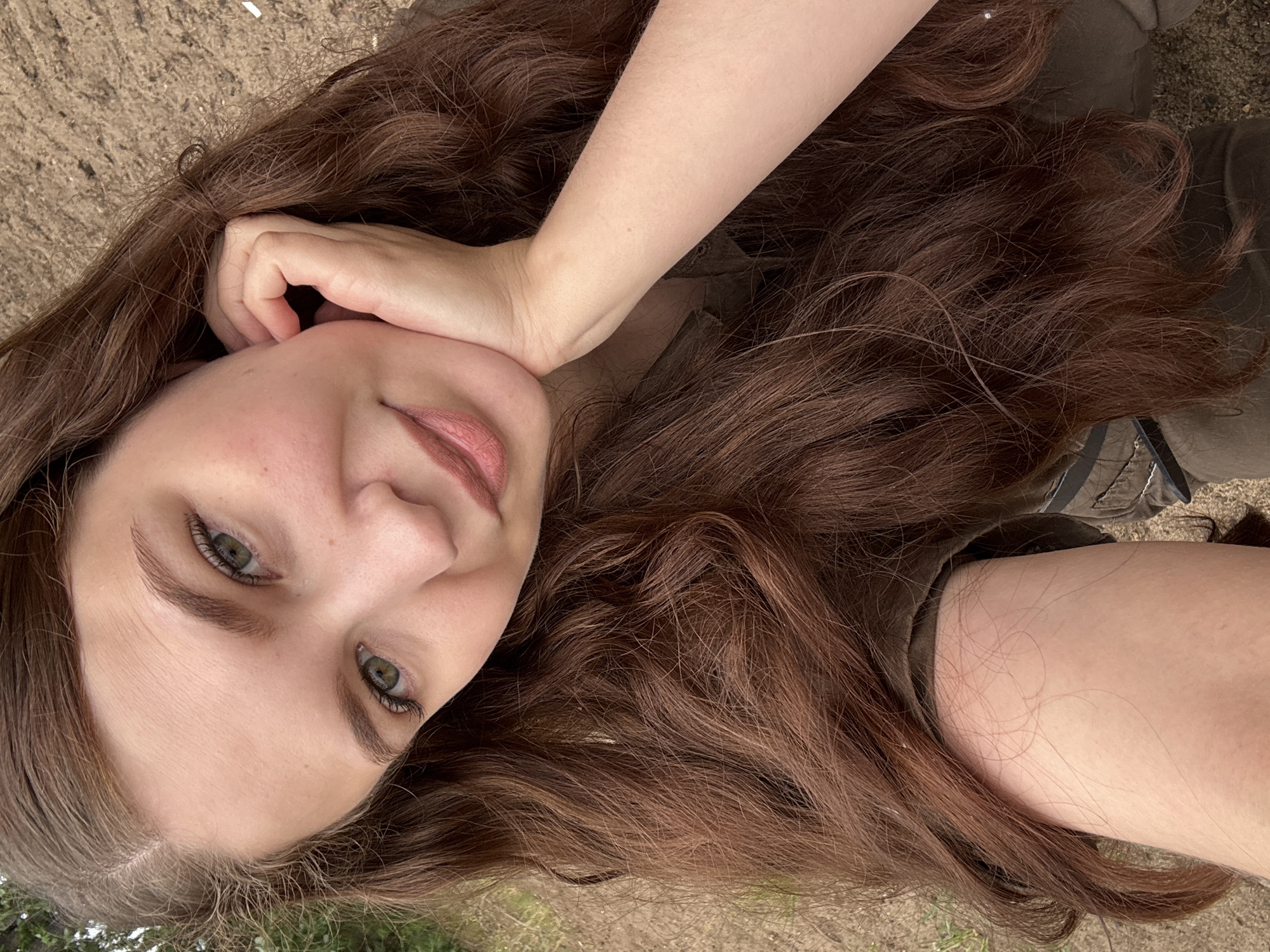This media file is a close-up, high-angle selfie of a young woman with long, wavy brown hair. She is lying down, with her head tilted and her right hand gently touching her cheek and resting near her temple. Her eyes are green and her lips are slightly parted, with a natural pinkish hue. Her expression appears relaxed and perhaps a bit contemplative.
The woman is wearing a dark, possibly olive-green, top. Her hair is the most prominent feature, cascading around her face and shoulders. The background is out of focus but appears to be an outdoor setting with earthy tones, suggesting a ground covered in dirt, sand, or dry leaves, and some blurred greenery in the lower portion of the image. The lighting suggests it is daytime, likely outdoors, with natural light illuminating her face. There are no visible text elements in the image. The overall mood is casual and intimate.