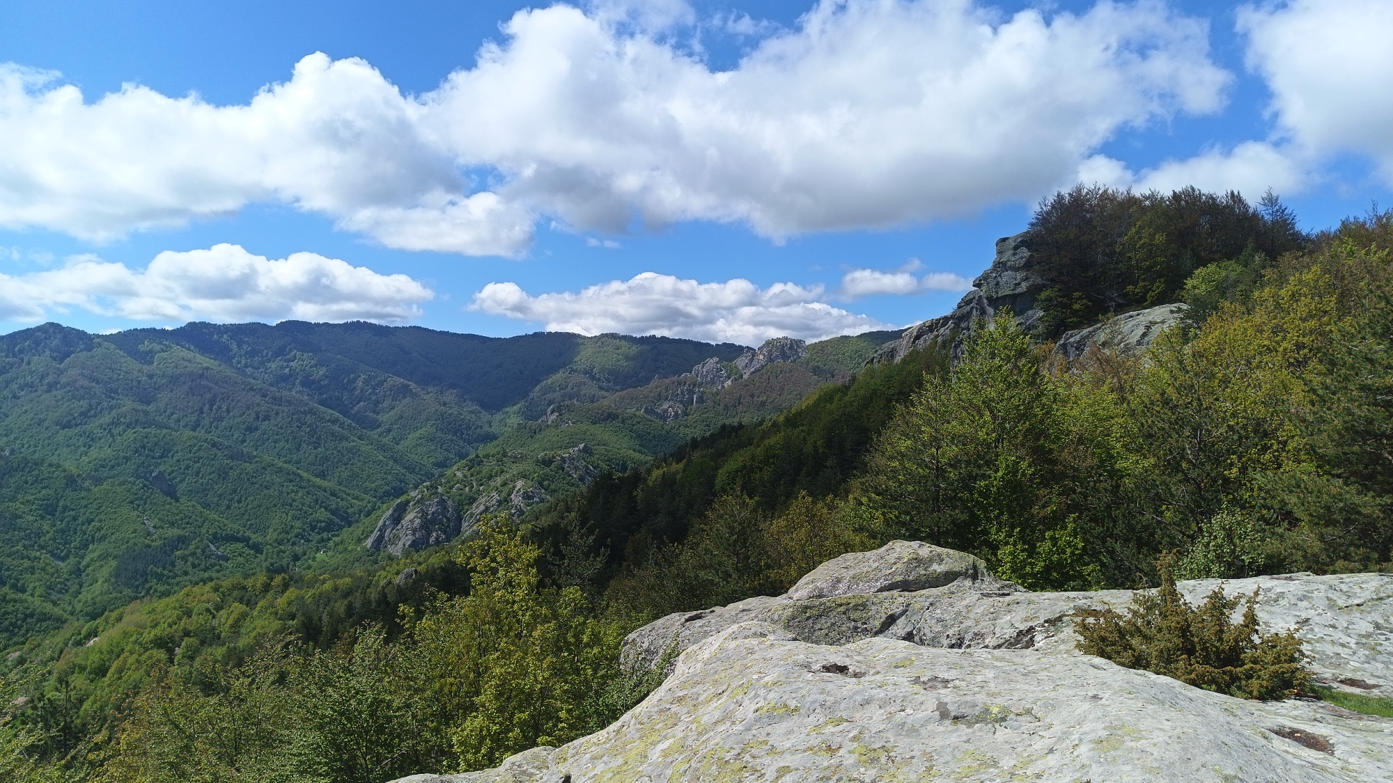 The image depicts a panoramic view of a mountainous landscape in the Sini Vrah area of Bulgaria. The foreground consists of large, lichen-covered rock formations. Green trees are visible at the foot of the rocks. In the mid-ground, a valley filled with dense forest stretches towards distant mountain ranges. The sky is blue with scattered white cumulus clouds.