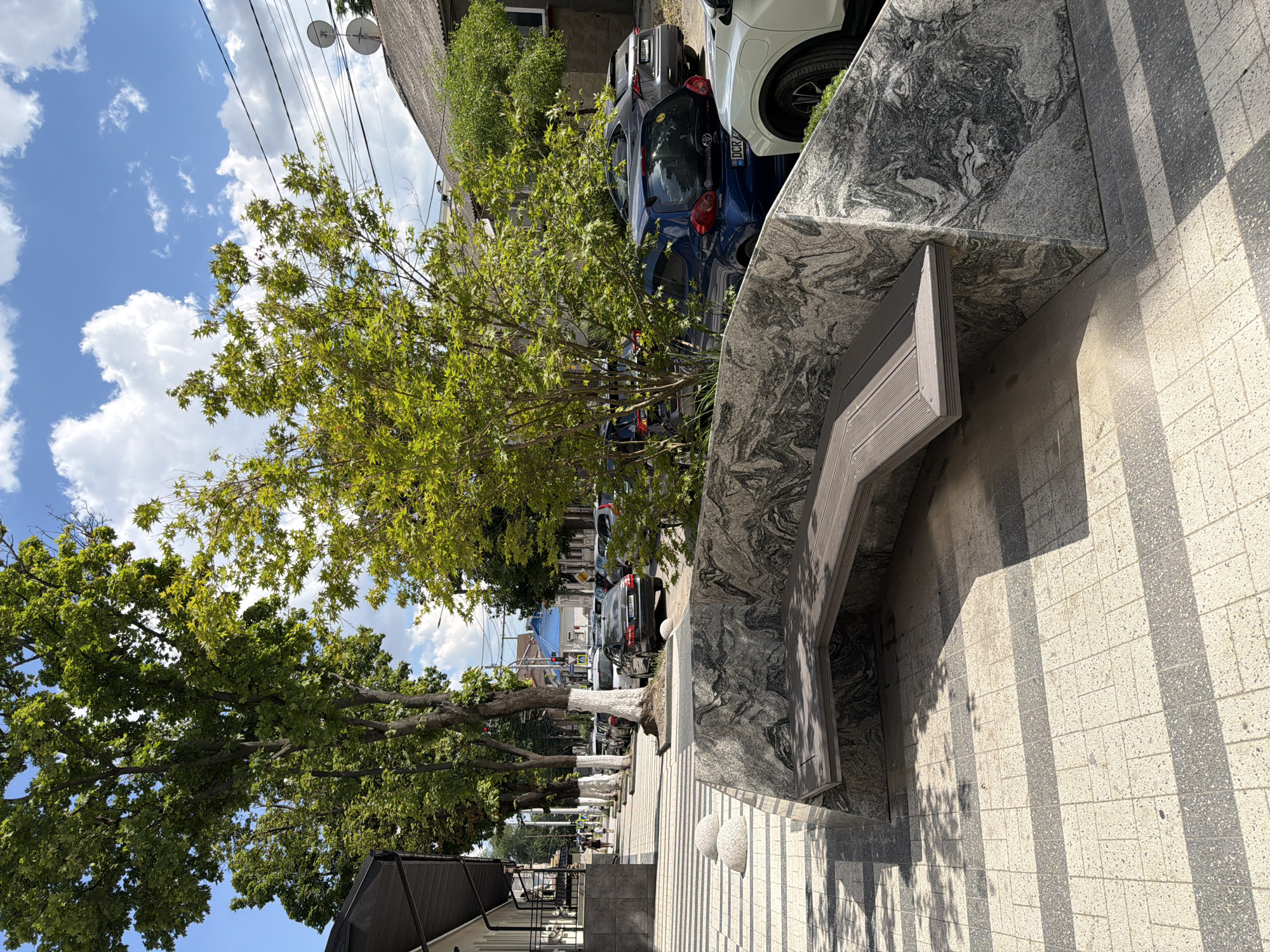 The image depicts a contemporary street scene in Chișinău, Moldova, featuring a modern granite bench integrated into the urban landscape. The bench, with its sleek, wooden seating, is positioned on a patterned pedestrian walkway composed of light-colored and dark gray tiles, creating a visually striking contrast. The walkway is bordered by a granite structure with a dramatic, swirled pattern, hinting at sophisticated urban design.
Behind the bench, a row of parked cars, including a blue Toyota and a white SUV, lines the street. Trees with lush green foliage cast dappled shadows on the walkway, adding a natural element to the scene. In the background, buildings with tiled roofs and various architectural styles are visible, alongside utility poles and wires, common elements of city infrastructure. The sky is a clear blue, dotted with fluffy white clouds, suggesting a pleasant day. A few individuals are present on the sidewalk, walking in the distance, indicating a functioning urban environment. The overall impression is one of a well-maintained public space designed for both aesthetics and functionality.