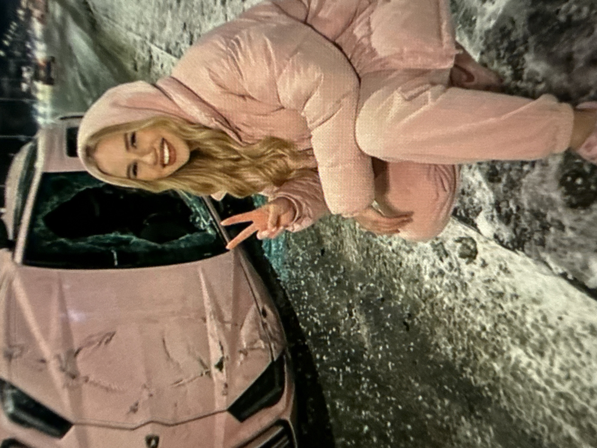 A young, fair-skinned woman with blonde hair, wearing a pink hooded jacket and matching sweatpants, kneels next to a pink Lamborghini car with a smashed windshield. The car also has damage to the front. She smiles and holds up two fingers in a peace sign. The ground is covered in what appears to be snow or ice. The location appears to be an urban environment at night.