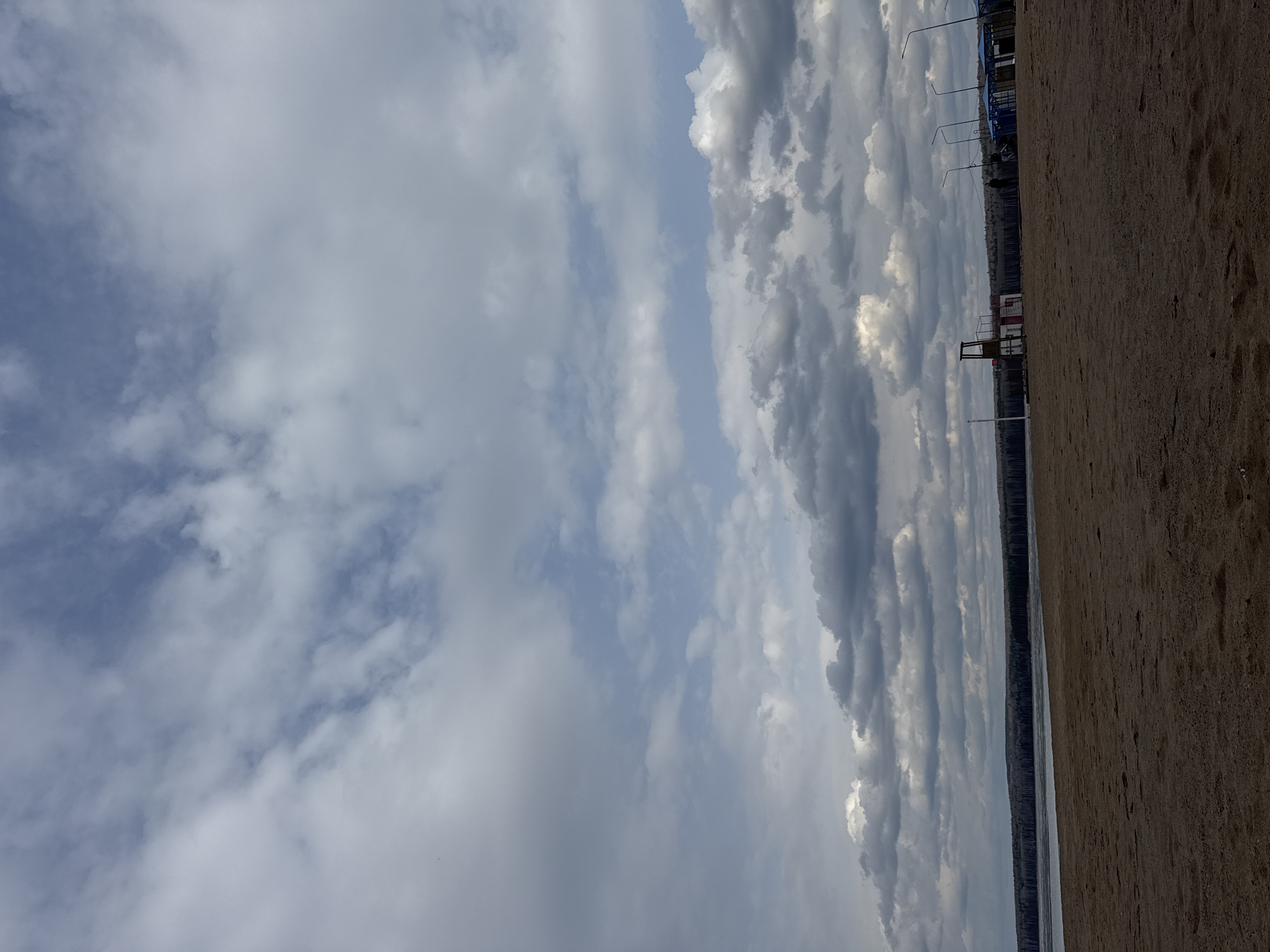The image shows a view from the beach in Chaikovsky, Russia. The scene is divided into thirds: the sky occupies the left third, filled with predominantly gray and white clouds. The middle third features a thin strip of water, followed by a line of trees, and then street lights and some buildings. The right third is sandy beach.