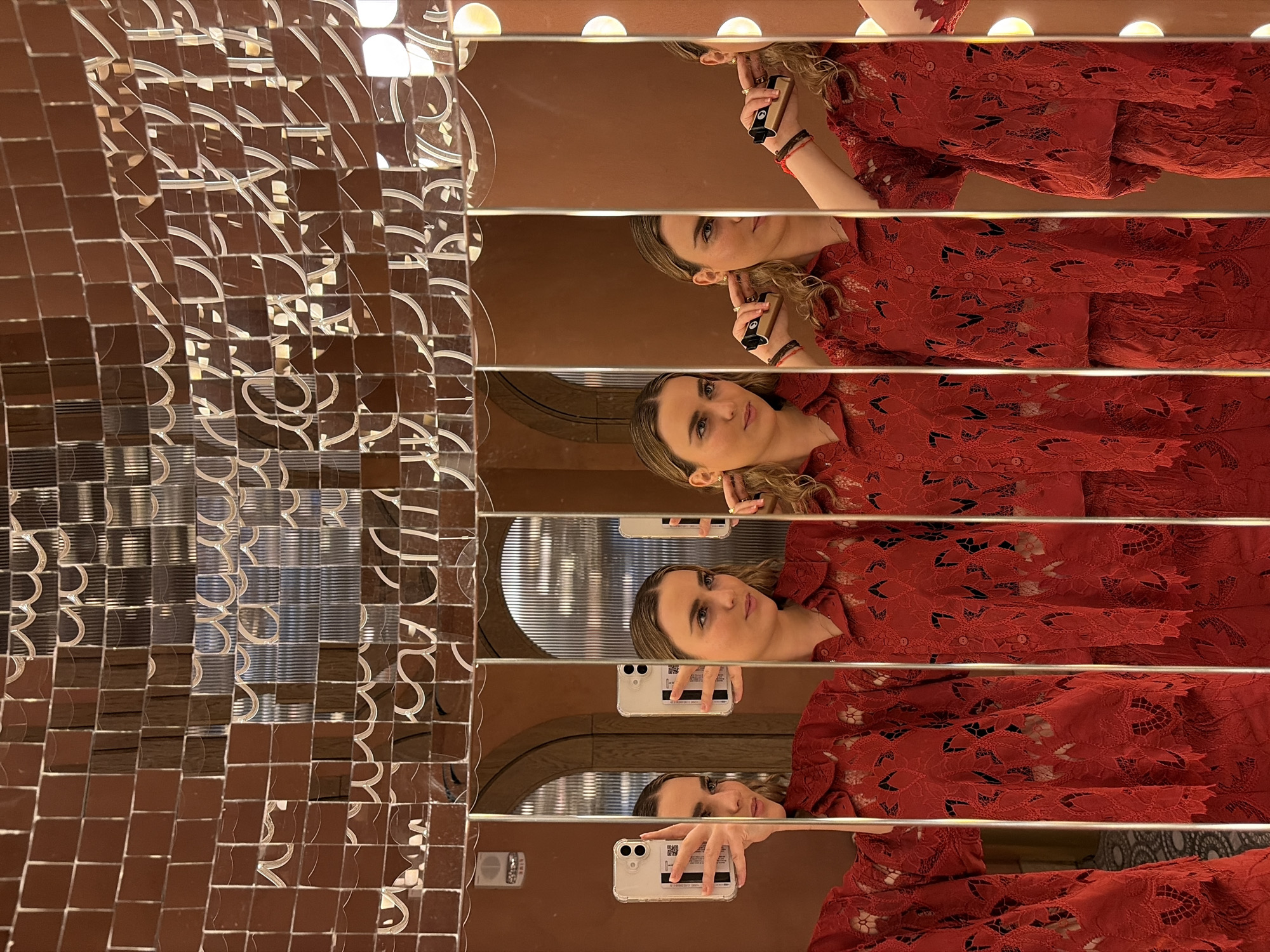 A woman in a red lace top holds a smartphone and takes a selfie in a mirror art installation. The installation features multiple horizontal mirrors, creating fragmented reflections of the woman and the surrounding environment. To the left of the mirrors is a wall covered in small, reflective brown tiles that catch reflections of lighting fixtures and the tiled wall. The scene is brightly lit, and the location is specified as Miami, United States of America.