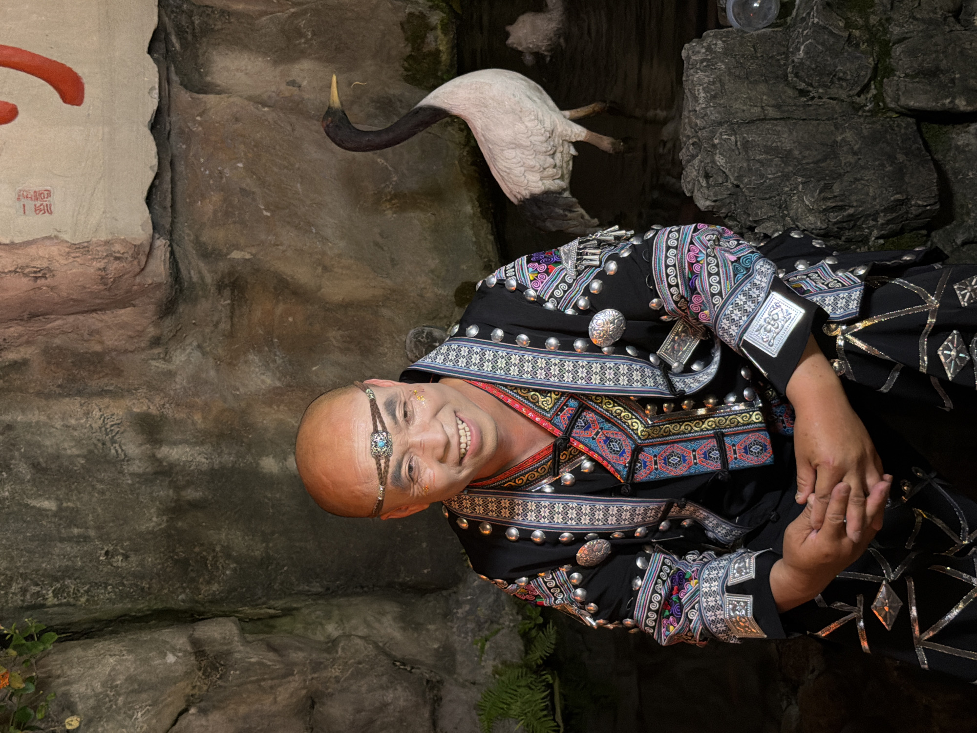 A middle-aged East Asian man with a shaved head and a wide smile is seated in what appears to be a performance setting. He is wearing an ornate black garment with intricate embroidery and silver embellishments, including a decorative headdress. His hands are clasped in his lap. Behind him, a lifelike prop of a crane stands in shallow water, with a rocky backdrop. The scene evokes a sense of cultural performance or storytelling. The image appears to have been taken in Yuzhong District, China.