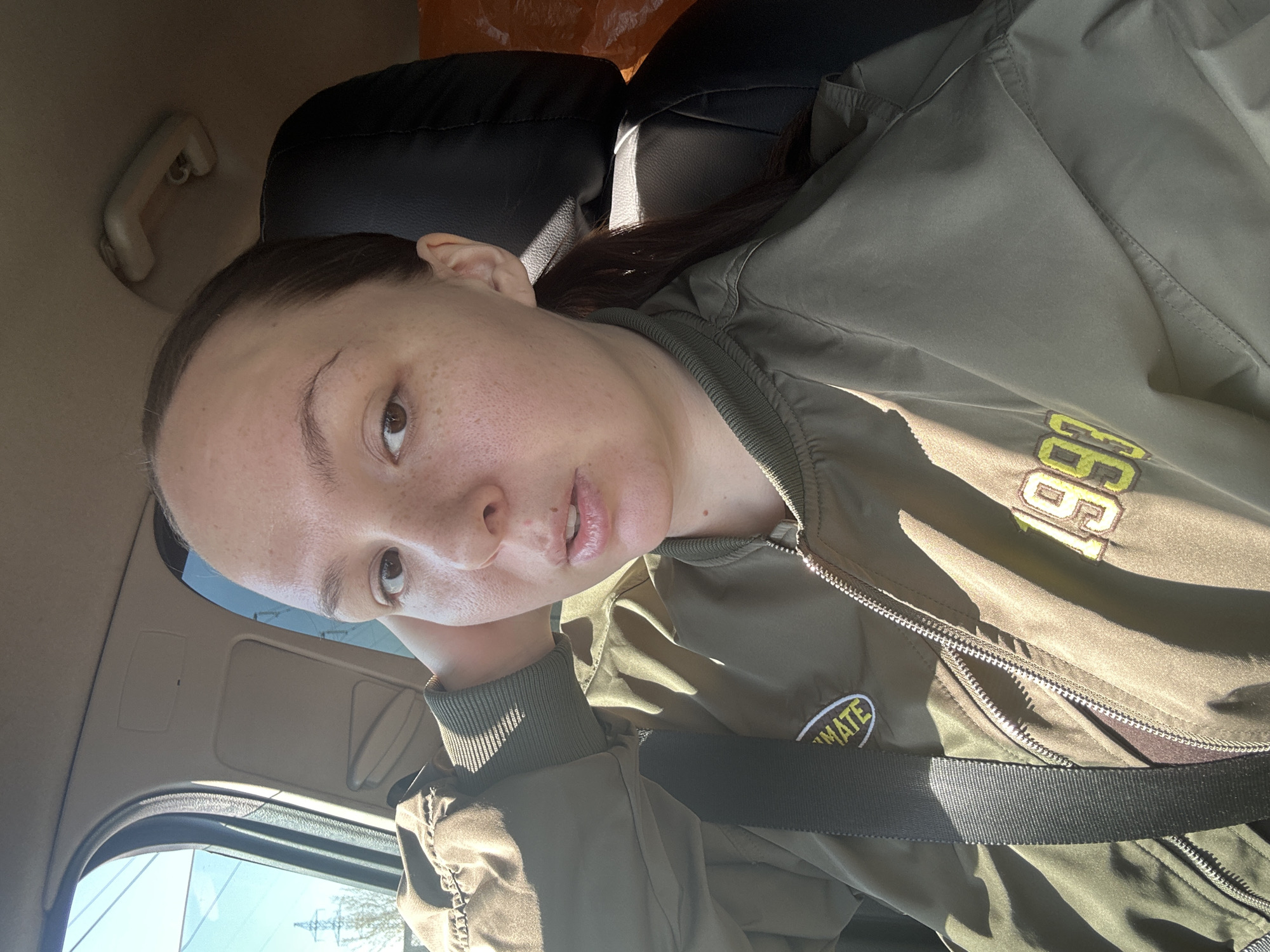 A woman is in the driver's seat of a car, wearing a green bomber jacket. The sun shines through the car window, creating a play of light and shadow on her face and jacket. The car's interior is beige and black. The location is presumed to be Chaikovsky, Russia based on the presence of buildings and foliage visible through the window.