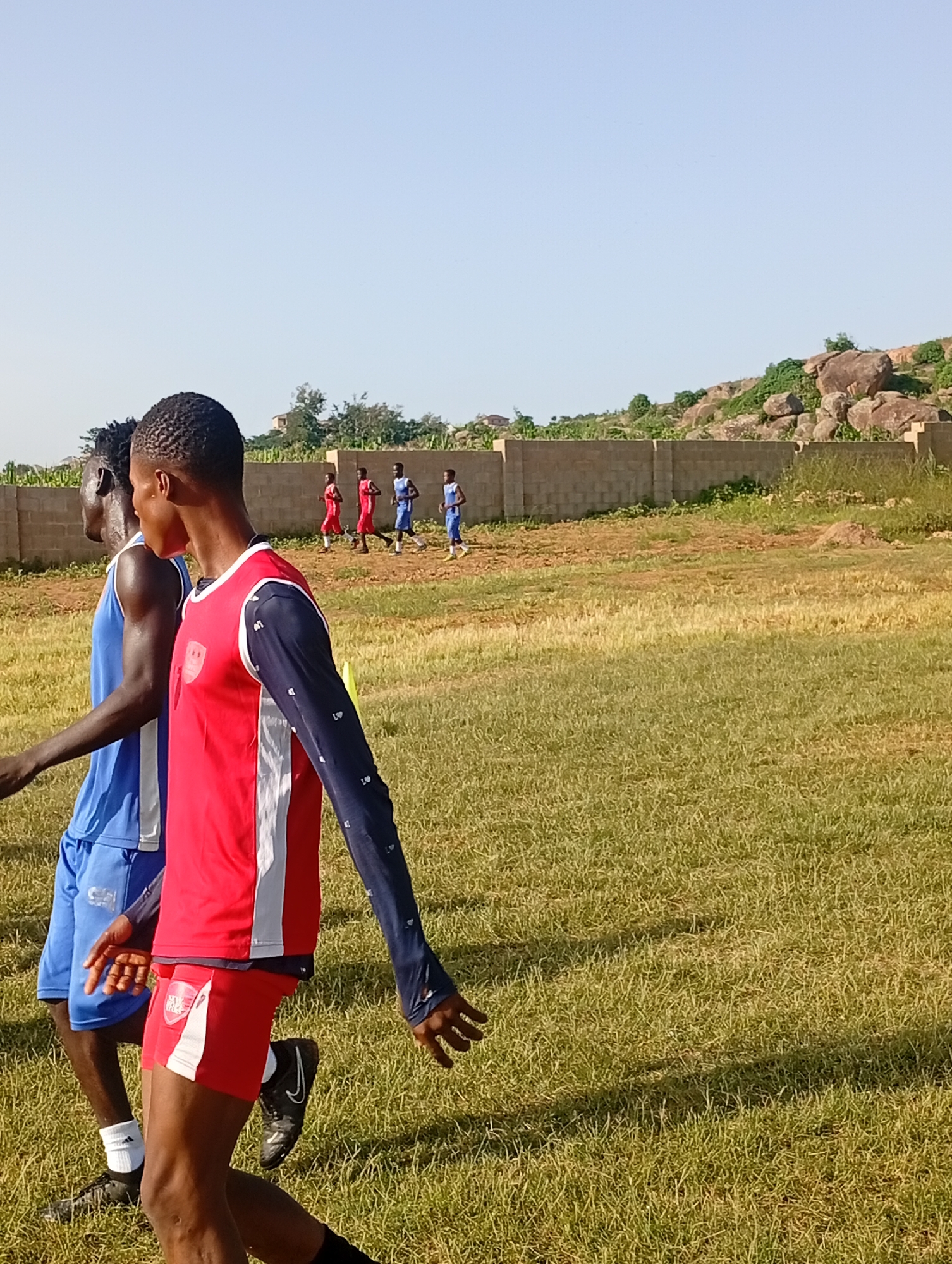 Two young men, appearing to be athletes, are walking or jogging across a grassy field. The man in the foreground on the right wears a red sleeveless shirt with a navy blue long-sleeved undershirt and red shorts with white trim. The man next to him on the left wears a blue sleeveless shirt and blue shorts. In the background, several other young men in similar athletic attire are visible, some wearing red shirts and others wearing blue shirts, running or walking further down the field. A low, light-colored brick wall runs horizontally across the middle ground of the image. Beyond the wall, a rocky, vegetated hillside rises against a clear, light blue sky. The ground is covered in dry, yellowish-green grass. The overall impression is a sports practice session in an outdoor, possibly rural or undeveloped, setting.