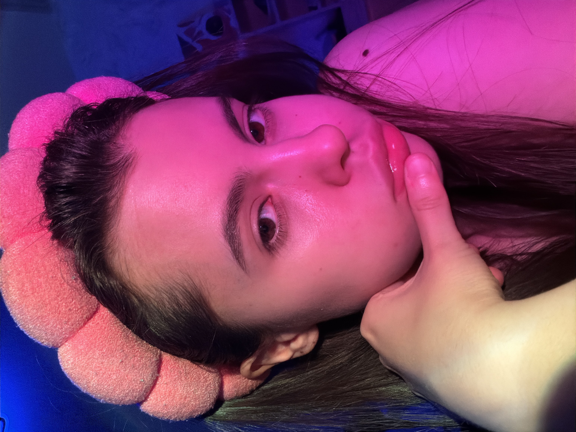 A young woman with long, dark hair is lying down, her head resting on a pink, flower-shaped pillow. Her face is illuminated by a pink light, while the background is cast in a blue light. Her right hand is raised to her face, her index finger touching her lip. The location, Chaikovsky, Russia, cannot be confirmed based on the image alone.