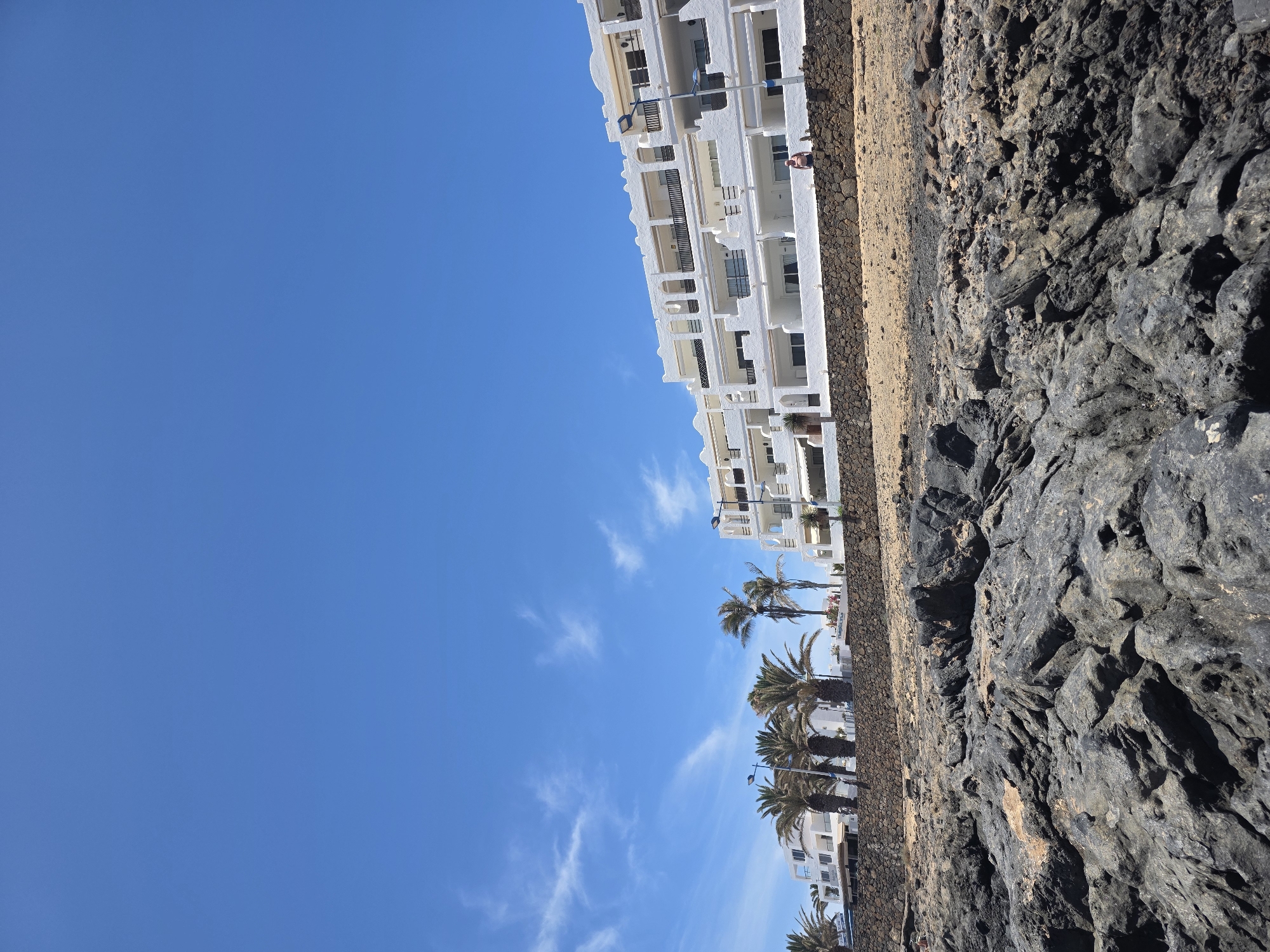 In Costa Teguise, Spain, a bright blue sky with wispy clouds forms the upper portion of the image. Below, a row of white apartment buildings with balconies and arched windows stretches across the horizon. Palm trees are interspersed between the buildings. In front of the buildings, a low stone wall separates them from a sandy embankment. In the foreground, dark, rugged rocks dominate the landscape, suggesting a coastal or volcanic environment. A person is visible sitting on the stone wall in front of the buildings, and another person is standing further to the right on the same wall. The scene depicts a sunny day at a seaside resort.