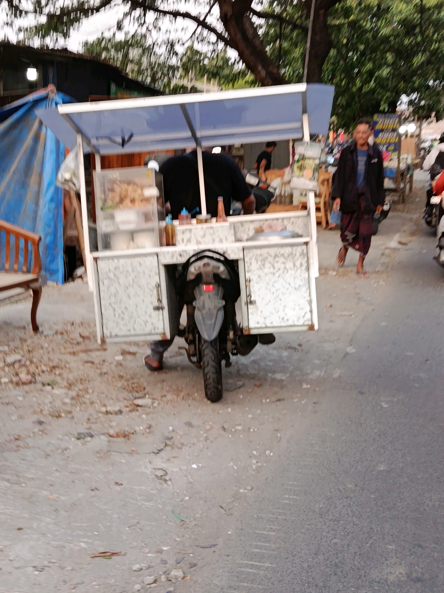 A vendor with a food cart attached to a motorcycle is parked on the side of a street in Bekasi, Indonesia. The cart is white with a blue canopy and displays food items in a clear container. A man, wearing a dark jacket and a patterned sarong, walks past the cart. Another person, wearing a black shirt, is visible behind the cart. The street appears to be made of asphalt, with debris scattered on the ground. Trees and other market stalls are visible in the background. The scene is illuminated by daylight, suggesting it is either morning or late afternoon.