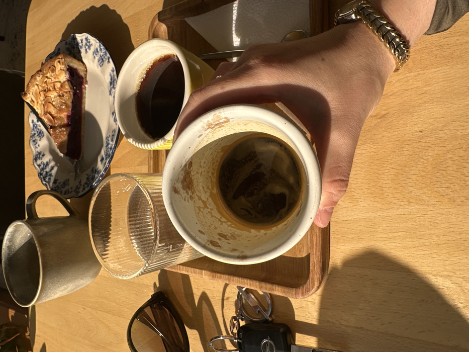 The scene depicts a light wooden tabletop in an establishment in Kazan, Russia, illuminated by direct sunlight that casts distinct shadows. A human left hand, wearing a gold-toned link bracelet watch, grips a white ceramic cup containing a small amount of dark liquid and visible interior residue. On a small wooden serving tray, a yellow ceramic cup holds dark coffee, alongside a folded white paper napkin and a partially visible metal spoon. Adjacent to the tray, a white ceramic plate with a blue floral pattern displays a single slice of berry pie topped with slivered almonds. A ribbed glass holds clear water, and a larger grey ceramic mug stands nearby. To the left of the tray, a pair of dark brown sunglasses and a keyring with a black car key fob, featuring an Opel logo, rest directly on the wooden surface.