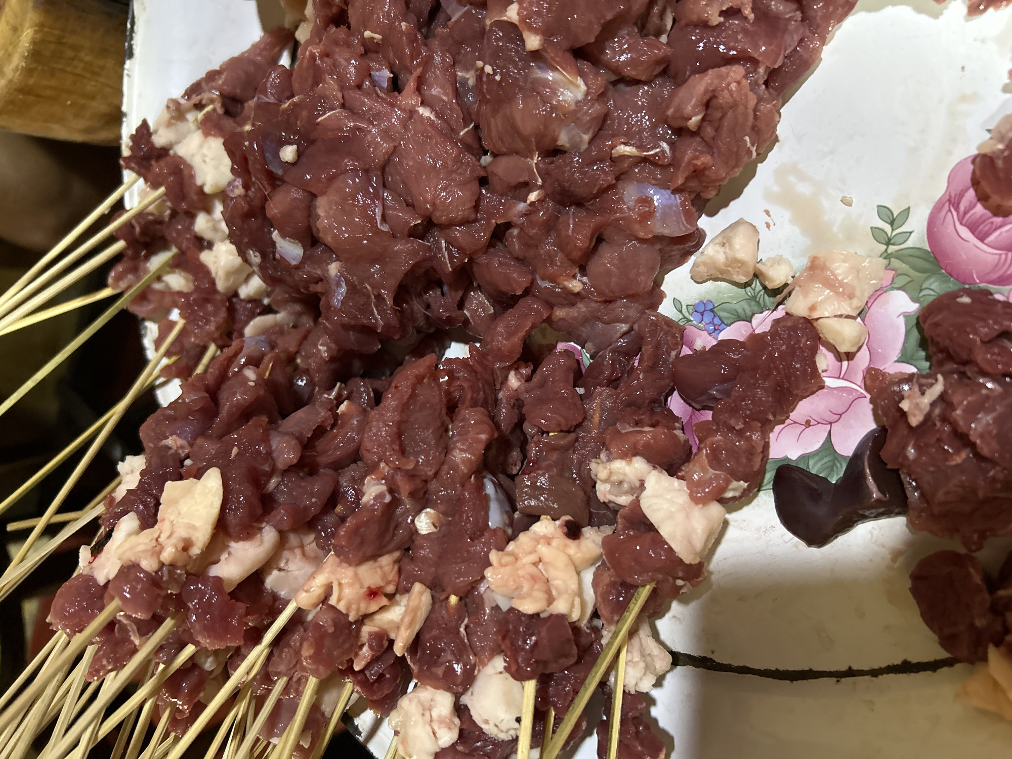 A close-up, overhead shot shows skewers of raw, cubed meat with some fatty pieces. The meat is reddish-brown and appears to be beef or lamb. The skewers are arranged on a light-colored platter with a floral pattern of pink and green. The platter has some visible liquid and small pieces of meat scattered on it. The background is dark and out of focus, with hints of wooden objects. The scene is consistent with food preparation in a home kitchen in Depok, Indonesia, likely for a meal such as satay.