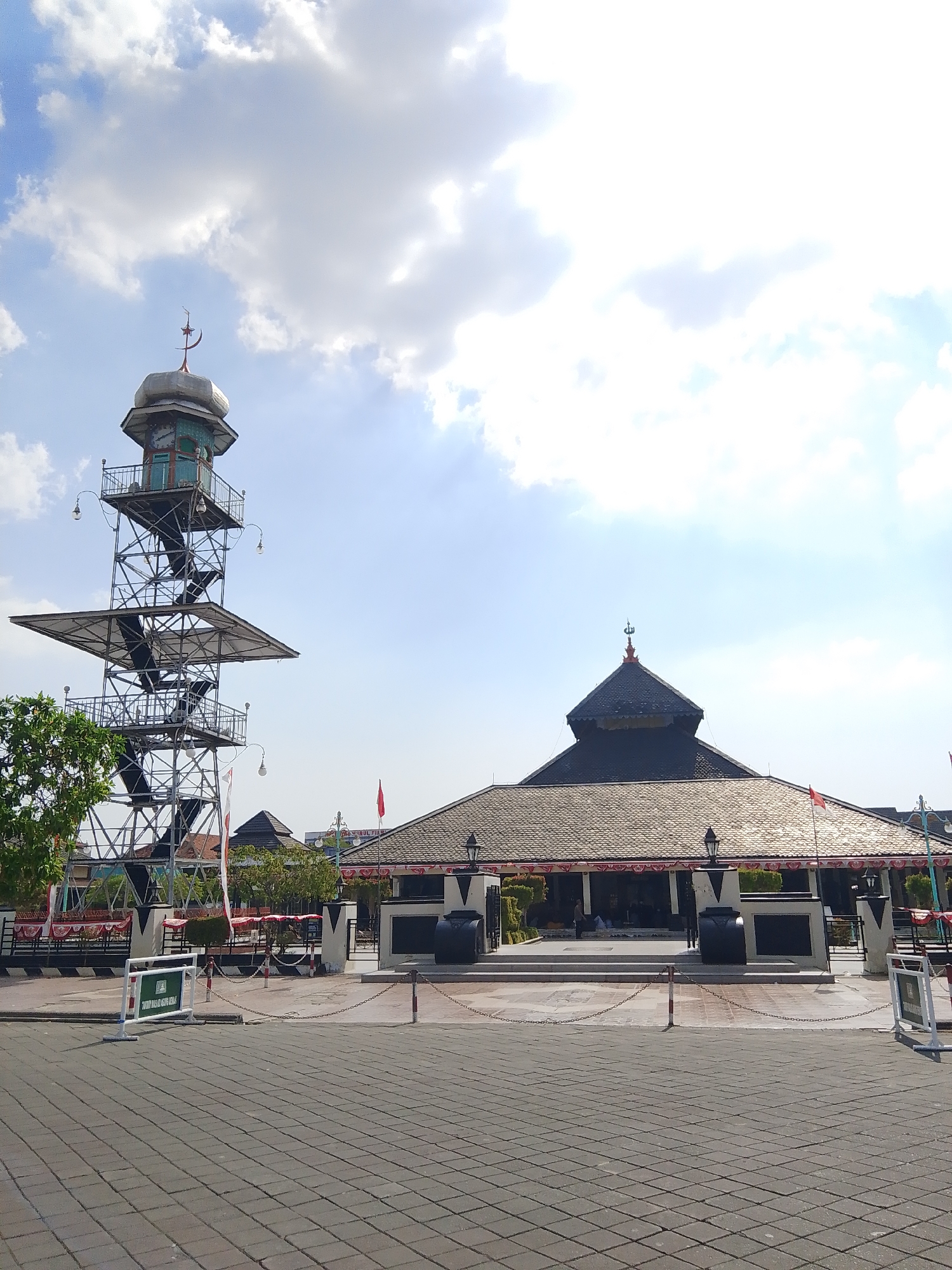 The image shows the Grand Mosque of Demak, a prominent historical mosque in the city of Demak, Indonesia. The mosque features a dark, tiered roof with ornate detailing and a spire topped with a crescent moon. Adjacent to the mosque is a tall, multi-level observation tower constructed of metal scaffolding. The tower has several platforms, a winding staircase, and a smaller structure at the top with a dome and a crescent. The foreground is paved with grey interlocking bricks, and chains on posts demarcate an area in front of the mosque. Red and white banners are visible along the mosque's facade. The sky is a bright blue with scattered white clouds, and sunlight is casting shadows. A tree with green foliage is partially visible on the left. Several people are present, some seated inside the mosque and others walking outside. A white sign with green text is placed on the paved area, and a red flag is flying near the mosque.