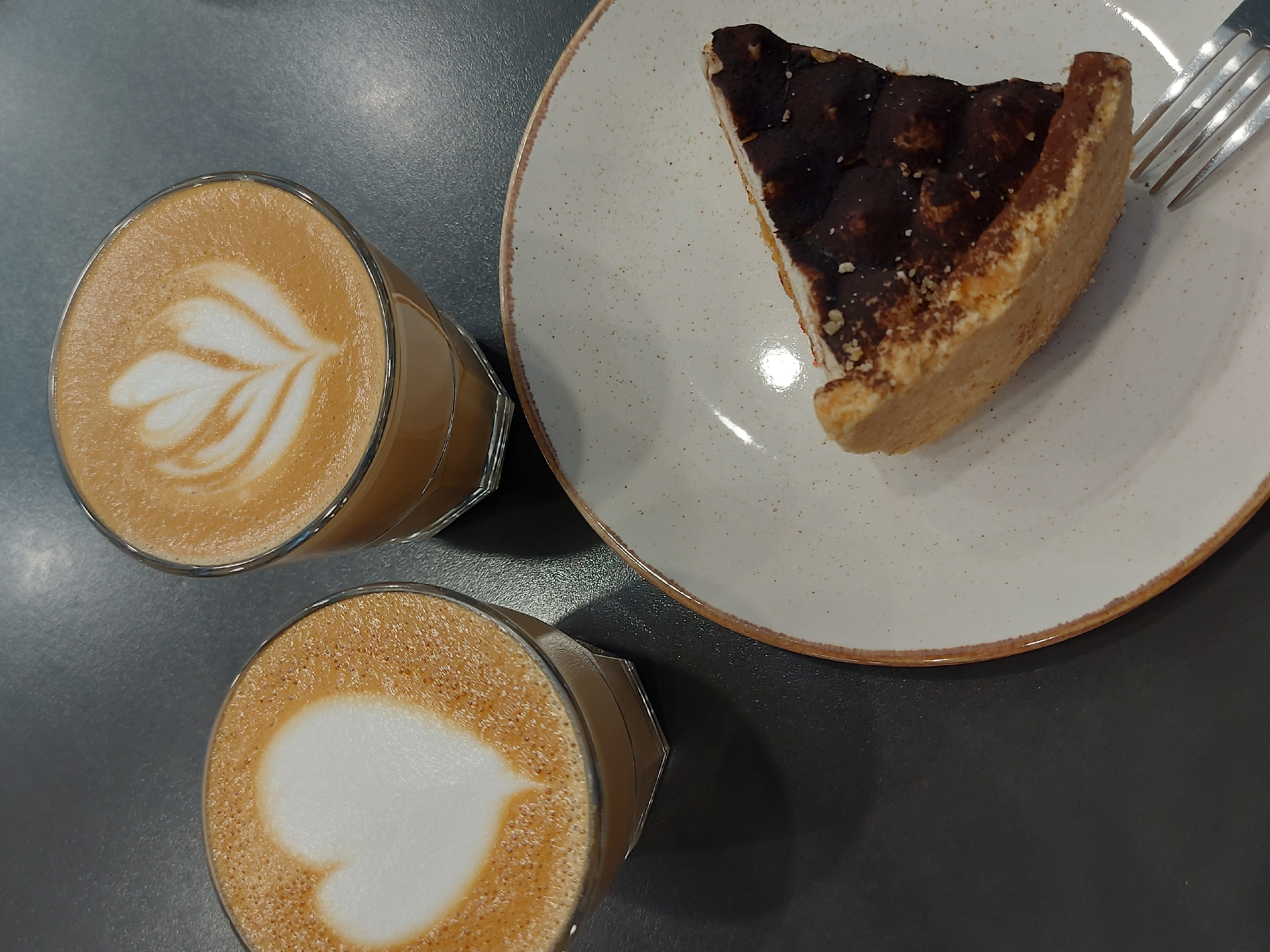 The image shows a close-up, overhead view of a table in a café, likely in Izhevsk, Russia, based on the origin of the image. Two glasses of latte with distinct latte art – one a heart shape and the other a leaf pattern – sit next to a plate containing a partially eaten slice of cake. A fork rests on the plate. The surface of the table is a solid, dark grey color.
