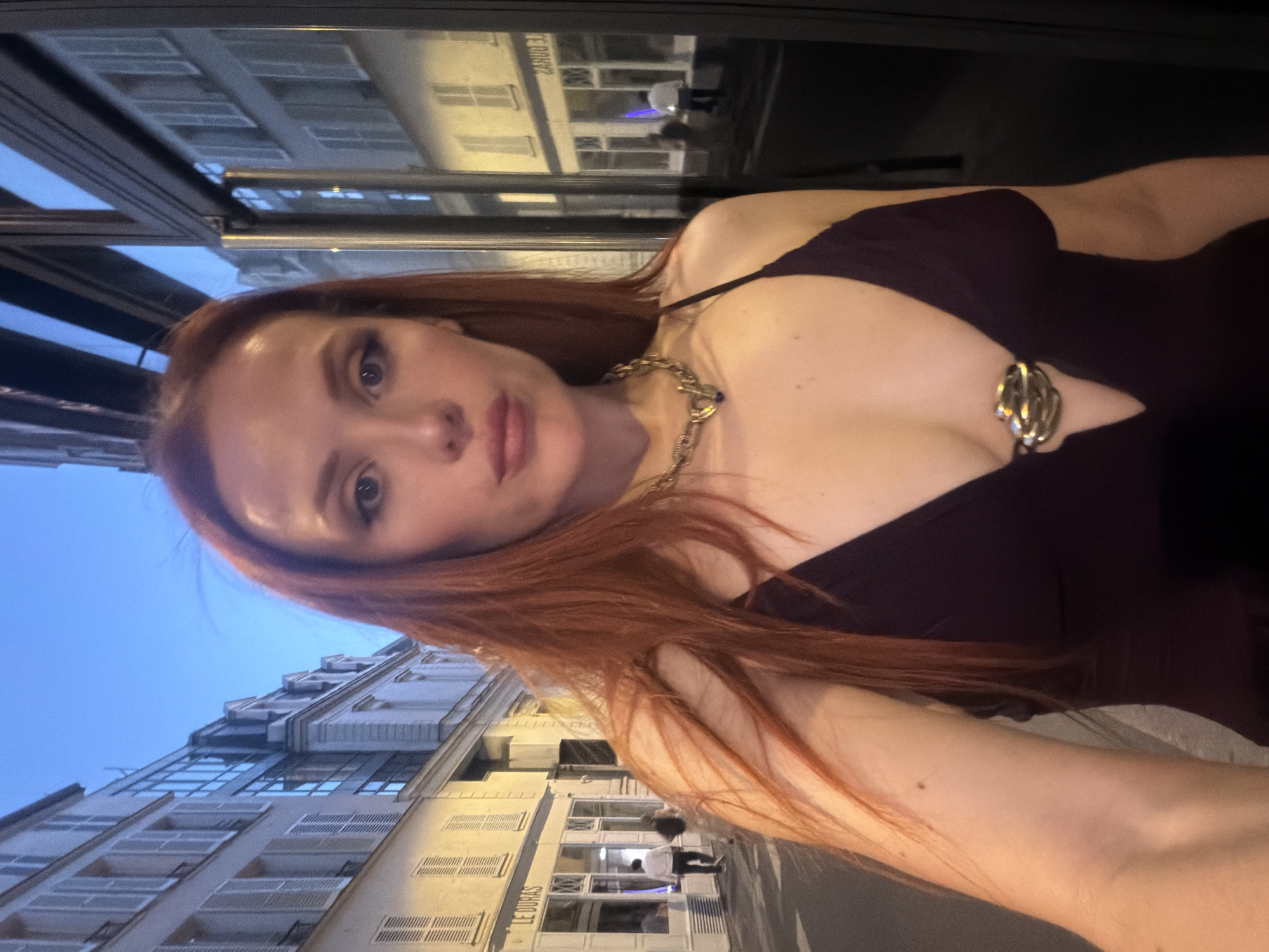 A woman with red hair is taking a selfie outdoors in Paris, France. She is wearing a low-cut, dark-colored dress and a gold necklace. The building behind her has a sign that reads "LE DURAS". Other buildings are visible, and the sky is blue. The photo is taken from a low angle.