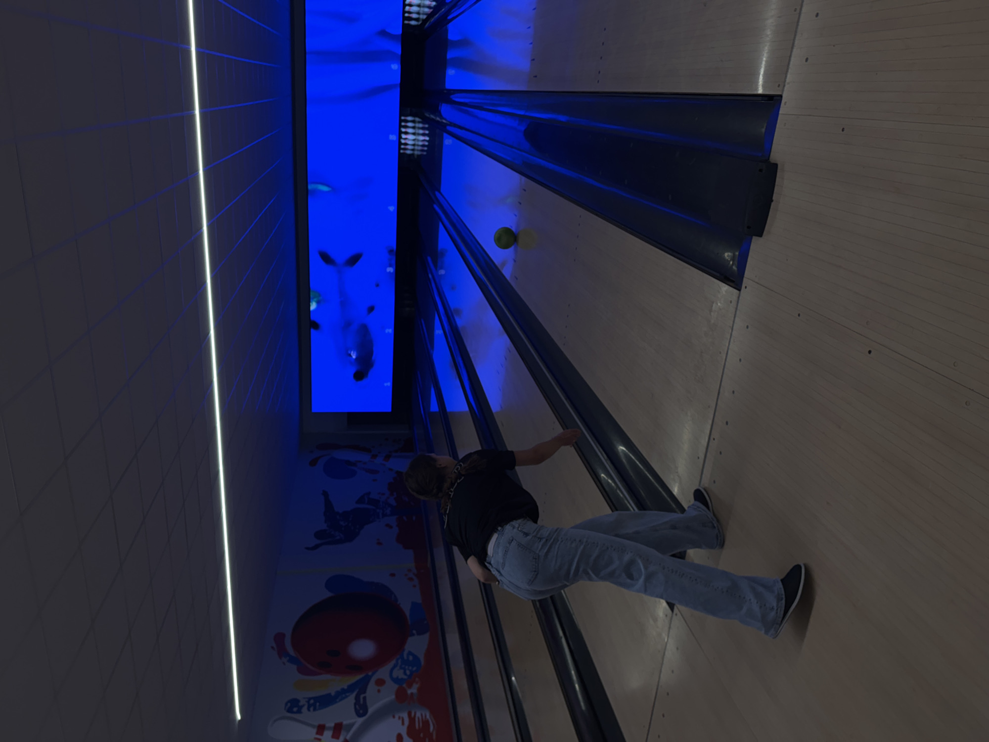 A person is bowling in an indoor bowling alley. The alley features blue painted gutters and a screen above with aquatic imagery. The person is wearing jeans, a black shirt, and dark shoes. A green bowling ball is in mid-roll down the lane. The location is possibly in Milan, Italy, but cannot be definitively determined from the image.
