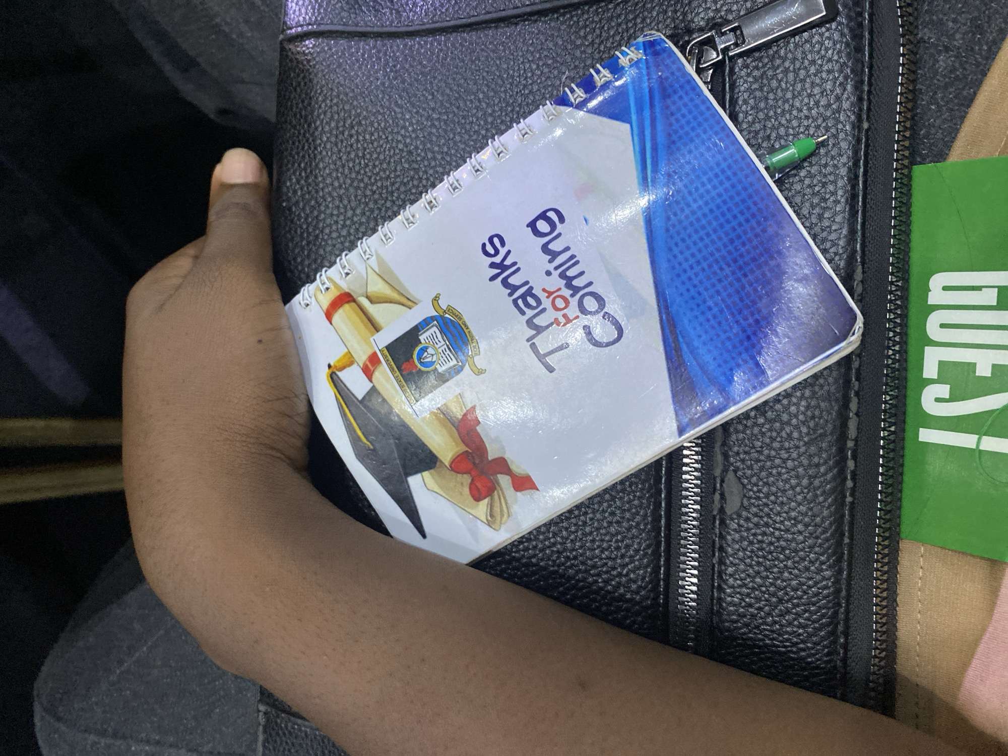 In Ikoyi, Nigeria, a close-up shot shows a person's hand resting on a black, textured bag. On the bag, a white spiral notebook is placed, its cover featuring a graduation cap, a diploma scroll with a red ribbon, and text that reads "Thanks For Coming." A small green pen is tucked into the notebook. To the left of the notebook, a green card with the word "GUEST" printed in white is visible. The background is dark, suggesting an indoor setting.