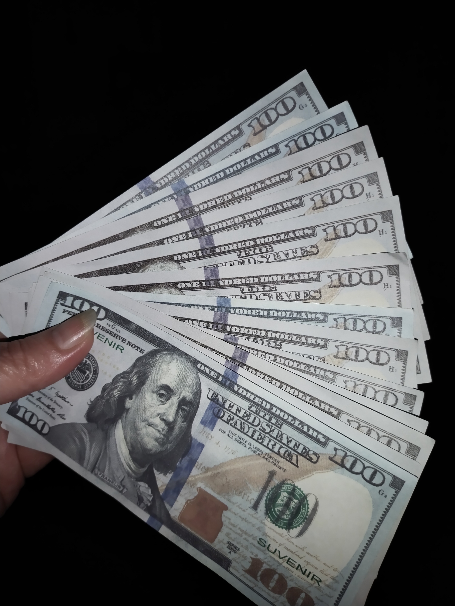 A hand is holding a fan of US $100 bills against a black background. The bills are partially visible and are spread out in a semi-circle. The image does not contain any information or context related to Horishni Plavni, Ukraine.