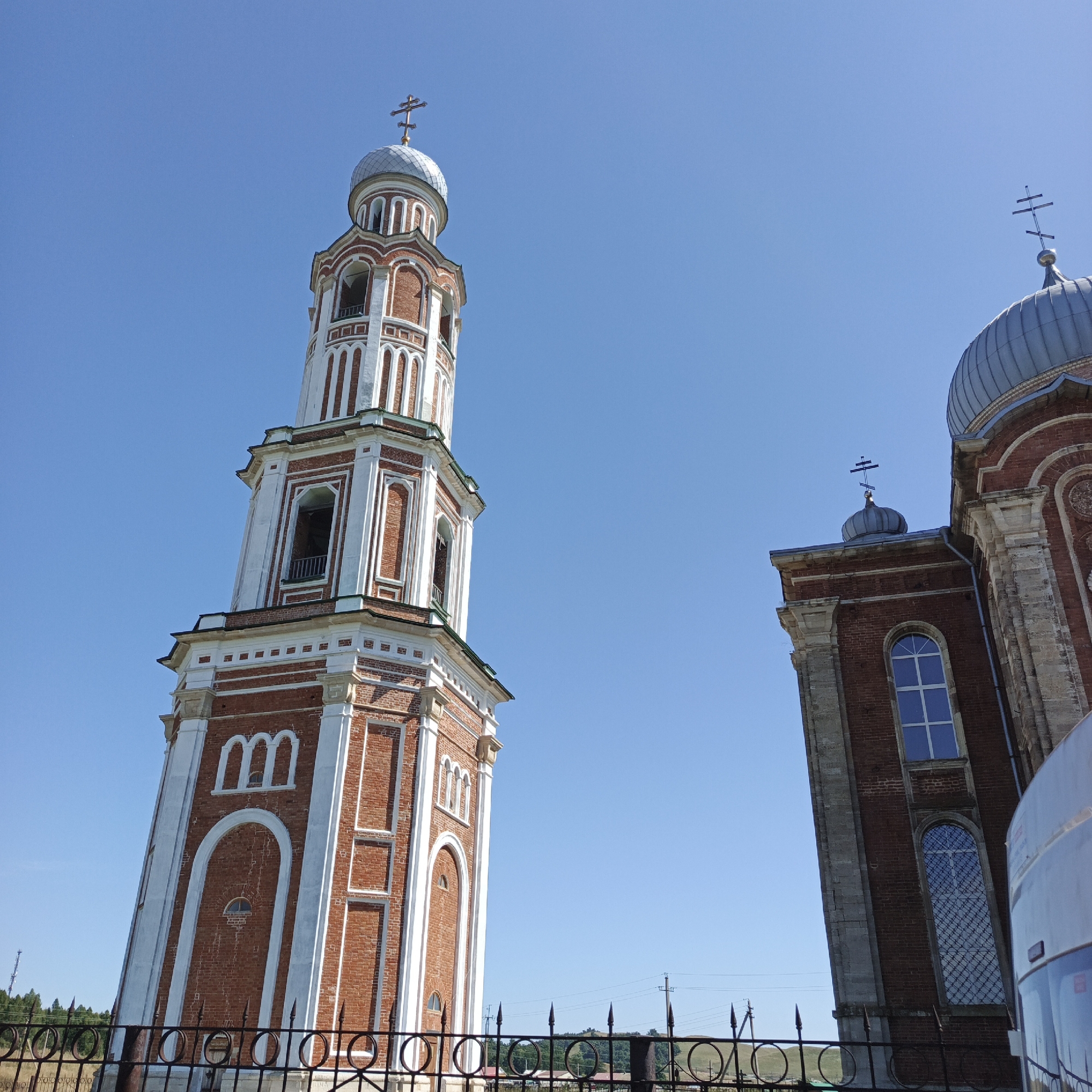 The image shows a brick bell tower and a portion of a church building against a clear blue sky. The bell tower, to the left, is tall and octagonal, with white decorative elements and arched openings. It is topped with a silver dome and a cross. To the right, a section of the church is visible, also constructed of brick with stone accents. It features a domed roof, a cross, and tall arched windows. A black metal fence runs along the bottom of the image, with a blurry background of green hills and some distant power lines. The overall scene depicts religious architecture under a bright sky.