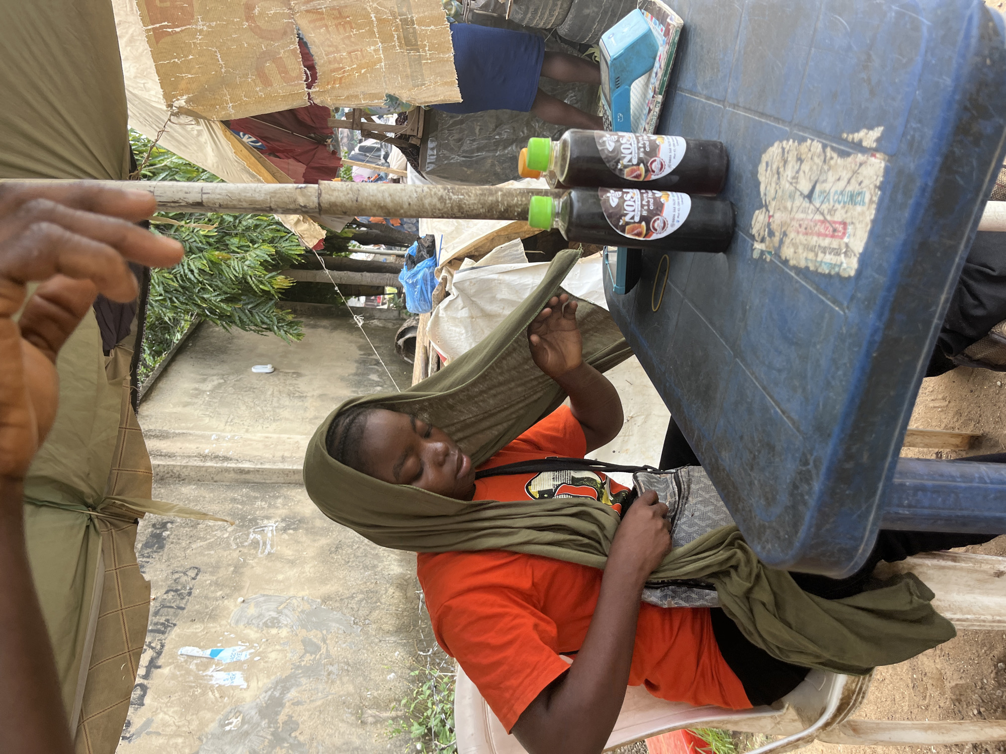 A young woman wearing an orange t-shirt and a green headscarf sits at a blue plastic table outdoors. She is holding a green cloth and appears to be adjusting it. On the table in front of her are two dark bottles with labels, a yellow rubber band, and a blue payment terminal. The table is set up in an outdoor market area, possibly in Abuja, Nigeria, as indicated by the environment. Behind her, there are other stalls and structures, suggesting a commercial setting. The background includes a textured wall and lush green trees. Another person's hand is visible in the upper part of the image, suggesting their presence nearby.