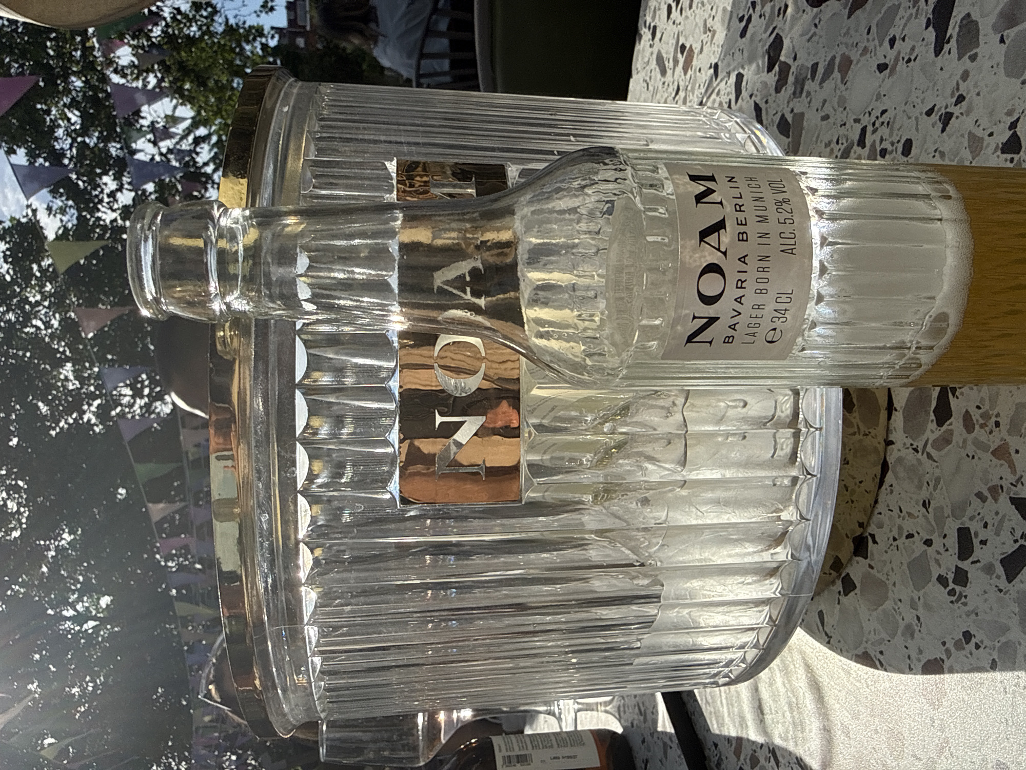 Here is a detailed description of the image:
The image shows a half-empty clear glass bottle with the label "NOAM BAVARIA BERLIN LAGER BORN IN MUNICH e 34CL ALC. 5.2% VOL" placed inside an ice bucket with a gold rim. The ice bucket is also made of clear glass with vertical ridges. The bottle is lying on its side inside the bucket, with some of the light-colored liquid and foam showing. The setting appears to be outdoors on a table with a speckled surface. In the background, out of focus, there are trees, a building, and what appears to be bunting. The location is likely a garden or patio area. The image provides no evidence to suggest that it is located in the city of London, United Kingdom.