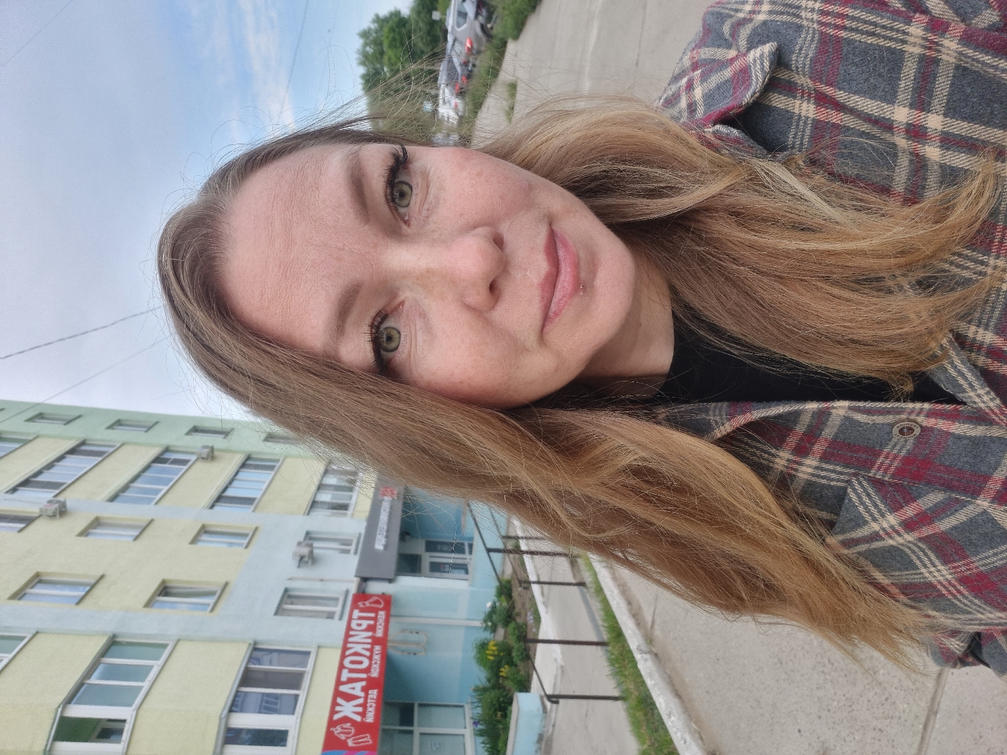 The image is a selfie of a young woman outdoors in Chaikovsky, Russia, captured from a slightly low angle. The woman, likely in her mid-20s to early 30s, has long, light brown hair and striking green eyes. She wears a multi-colored plaid shirt, predominantly grey with red and off-white lines, over a dark undershirt. A small, silver labret piercing is visible on her lower lip. She looks directly at the camera with a gentle smile and a calm, friendly expression.
The background shows an urban environment. Behind the woman is a multi-story building painted in light yellow-green and blue sections, featuring numerous windows. A prominent red sign with mirrored Cyrillic text is attached to the building. The main mirrored text appears to be "ТЬЯНТОЖАТ", with smaller, less legible Cyrillic words below it, including what looks like "МАКСОН" (MAXON) and "ЯДЕЛЬСКИЙ". Beyond the building, a street or paved area is visible, with parked cars and trees in the distance. The sky is bright blue with some white clouds, suggesting it is daytime with soft, diffused lighting.