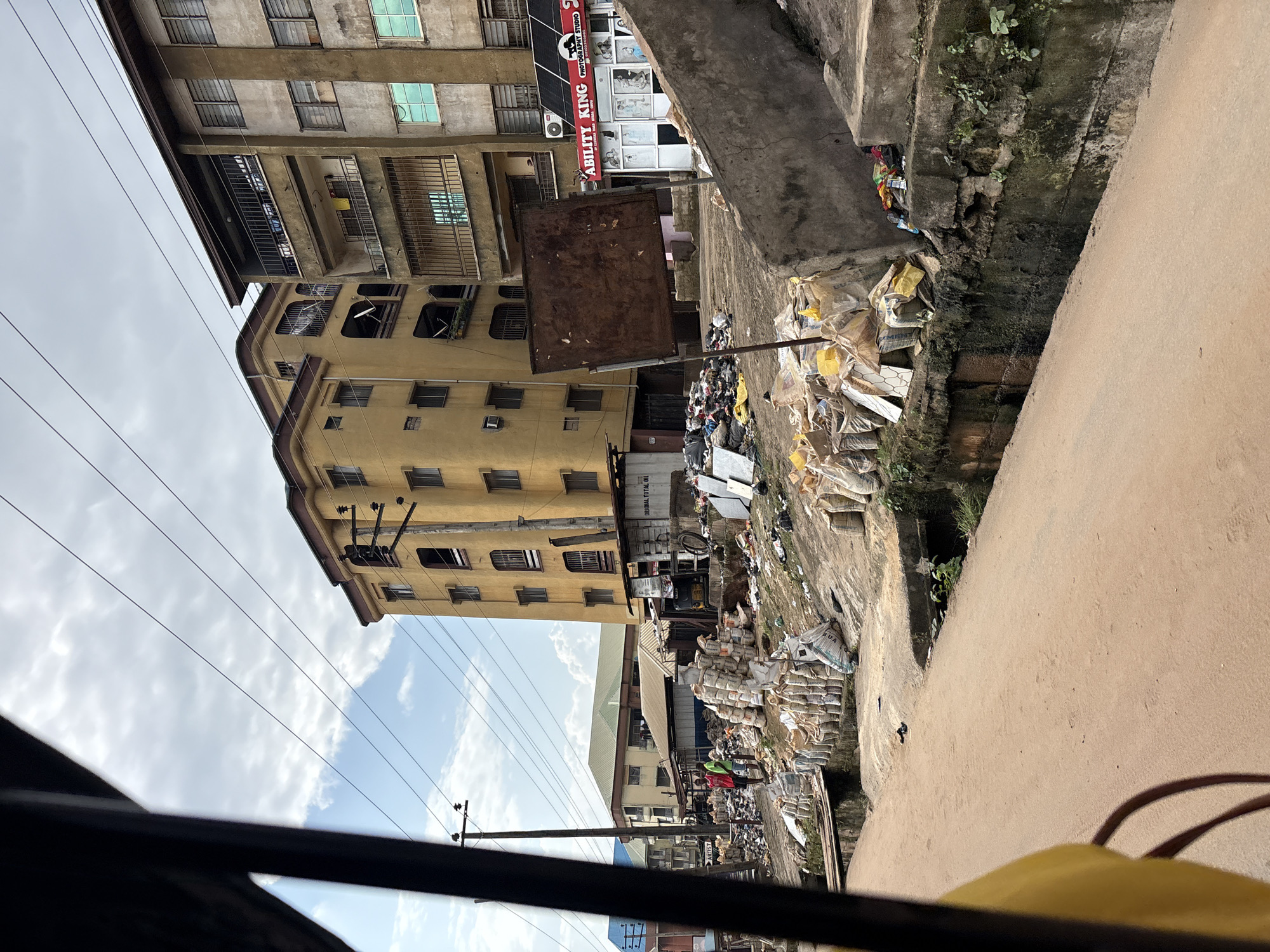 The image depicts a street scene in Onitsha, Nigeria. In the foreground, a sandy, unpaved road runs diagonally from the bottom left to the bottom center. To the right of the road is a concrete embankment, partially overgrown with vegetation, and a drainage ditch. Piled along the embankment are numerous sacks, many of which appear to be filled with goods, possibly cement or sand, and some loose debris.
Further back, there is a collection of various structures. A prominent yellow building with multiple floors and balconies stands to the right. Adjacent to it is a taller, grayer building with several stories, also featuring balconies. In front of these buildings, there are smaller, single-story structures, some with corrugated metal roofs, and what appears to be an open-air market or storage area.
A rusty, square signpost stands in the middle ground. Across the street, beyond the embankment and sacks, are more buildings and what seems to be a shop with a sign that reads "ABILITY KING PHOTOGRAPHY". Scattered around the area is a significant amount of litter and discarded materials, including plastic bags and other refuse, suggesting a general lack of waste management. Two individuals, men, are visible standing near the piled sacks, one wearing a red shirt and the other a green shirt. Power lines crisscross the sky, and the weather appears overcast with some clouds. The overall impression is one of a busy, somewhat unkempt urban environment.