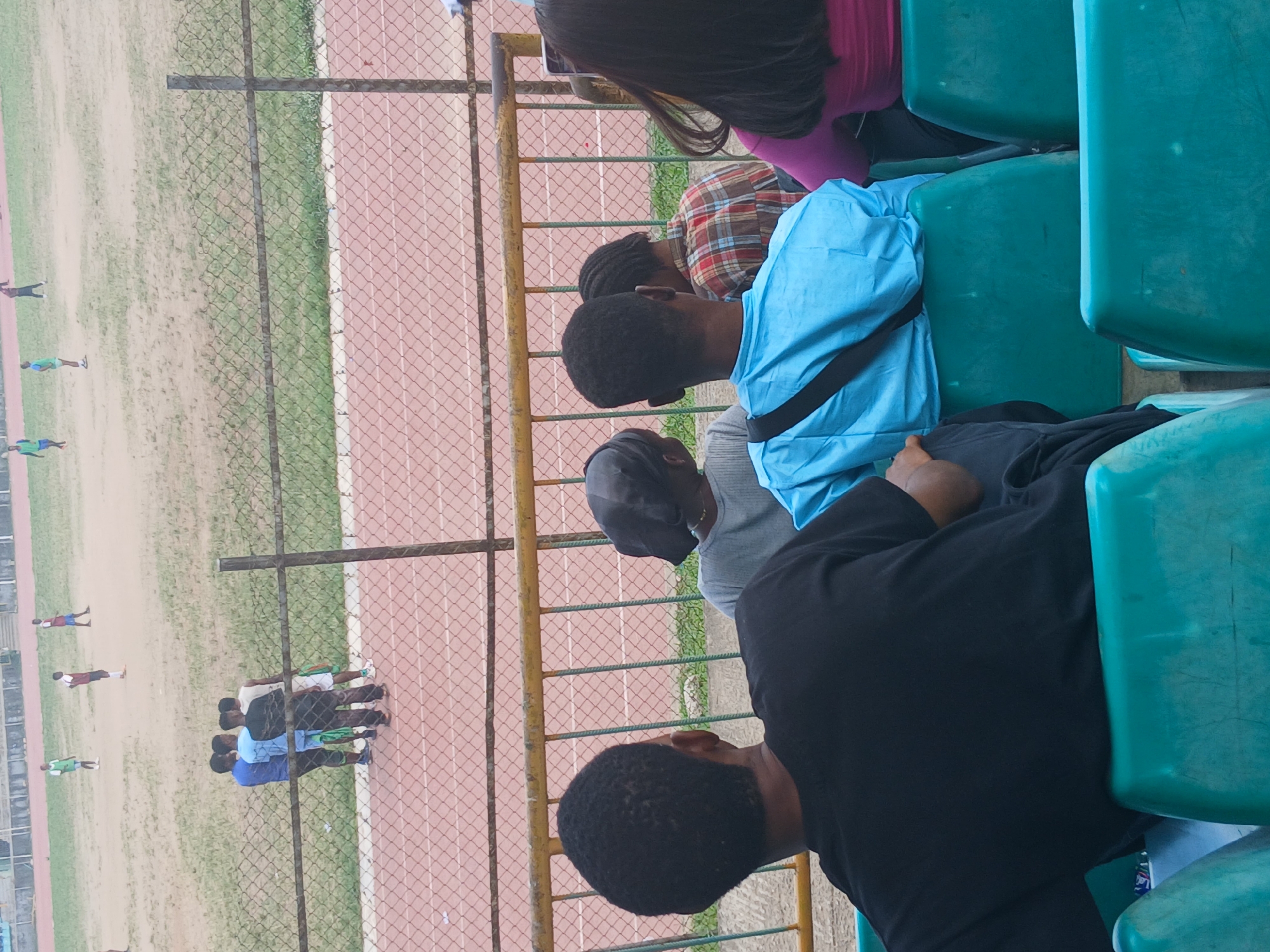The image depicts a group of people sitting in green stadium seats, observing activities on a field beyond a fence. The field includes a running track and a grassy area. Some individuals in athletic attire are visible on the field. The presence of a running track suggests the location is a sports stadium or athletic facility, likely in Lagos, Nigeria, given the context. The people in the stadium seats appear to be spectators or observers of the activities on the field. The fence separates the seating area from the field of play.