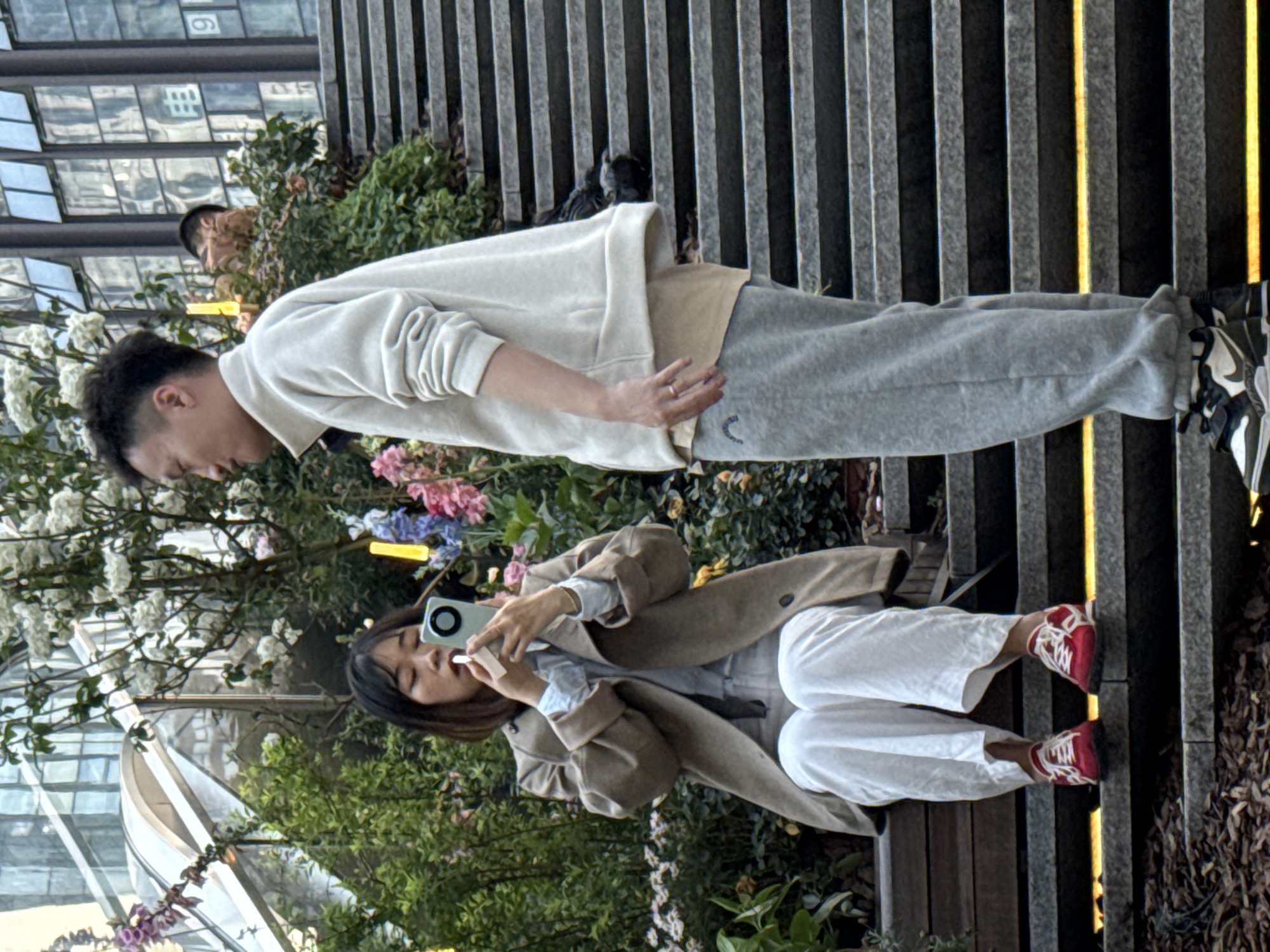 A man and woman are photographed at an unknown location within Liangjiang, China. The woman is seated on a bench, holding a smartphone in both hands, facing the camera, and is wearing a tan jacket, light blue shirt, white pants, and red sneakers. The man is standing and facing away from the camera, wearing a light-colored sweatshirt and gray sweatpants. The background contains greenery, flowers, a building with glass windows, and a partially obscured person. Additionally, there is a gray vertical-striped background with yellow lighting.