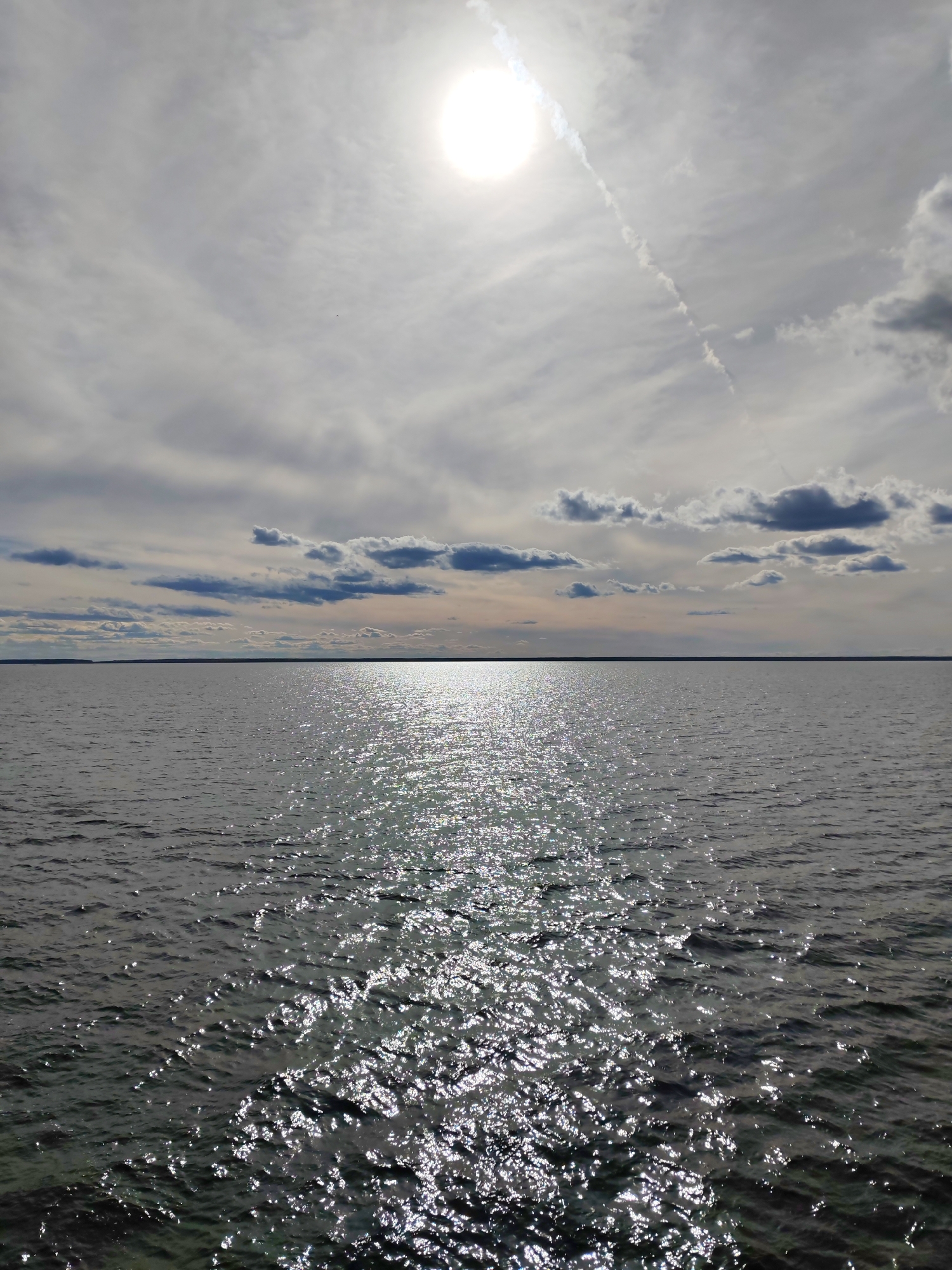 The image depicts a wide body of water, possibly a lake or sea, under a partly cloudy sky. The sun is visible, high in the sky, creating a path of reflected light on the water's surface. The water appears to have small waves causing the reflection to scatter and shimmer. A distant shoreline is visible, marked by a dark line, suggesting a landmass on the horizon. A thin contrail stretches from the sun and disappears into the clouds. The location is Gorushka, Russia.