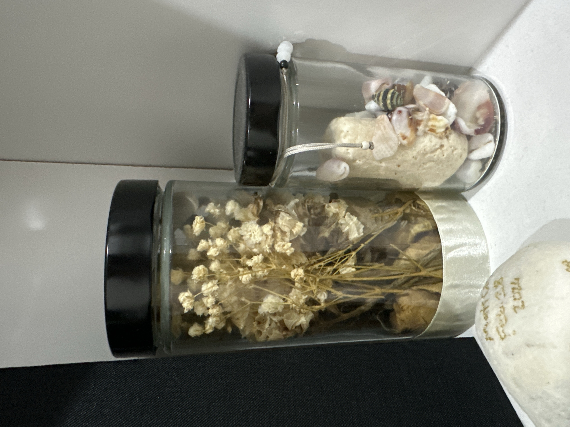 Two cylindrical glass jars with black lids sit atop a dark, possibly black fabric surface. The jar in the foreground contains dried, light-colored flowers. The jar in the background contains a rock and several seashells. The environment appears to be inside a room with a white shelf above the jars. The image was taken in Davao City, Philippines.