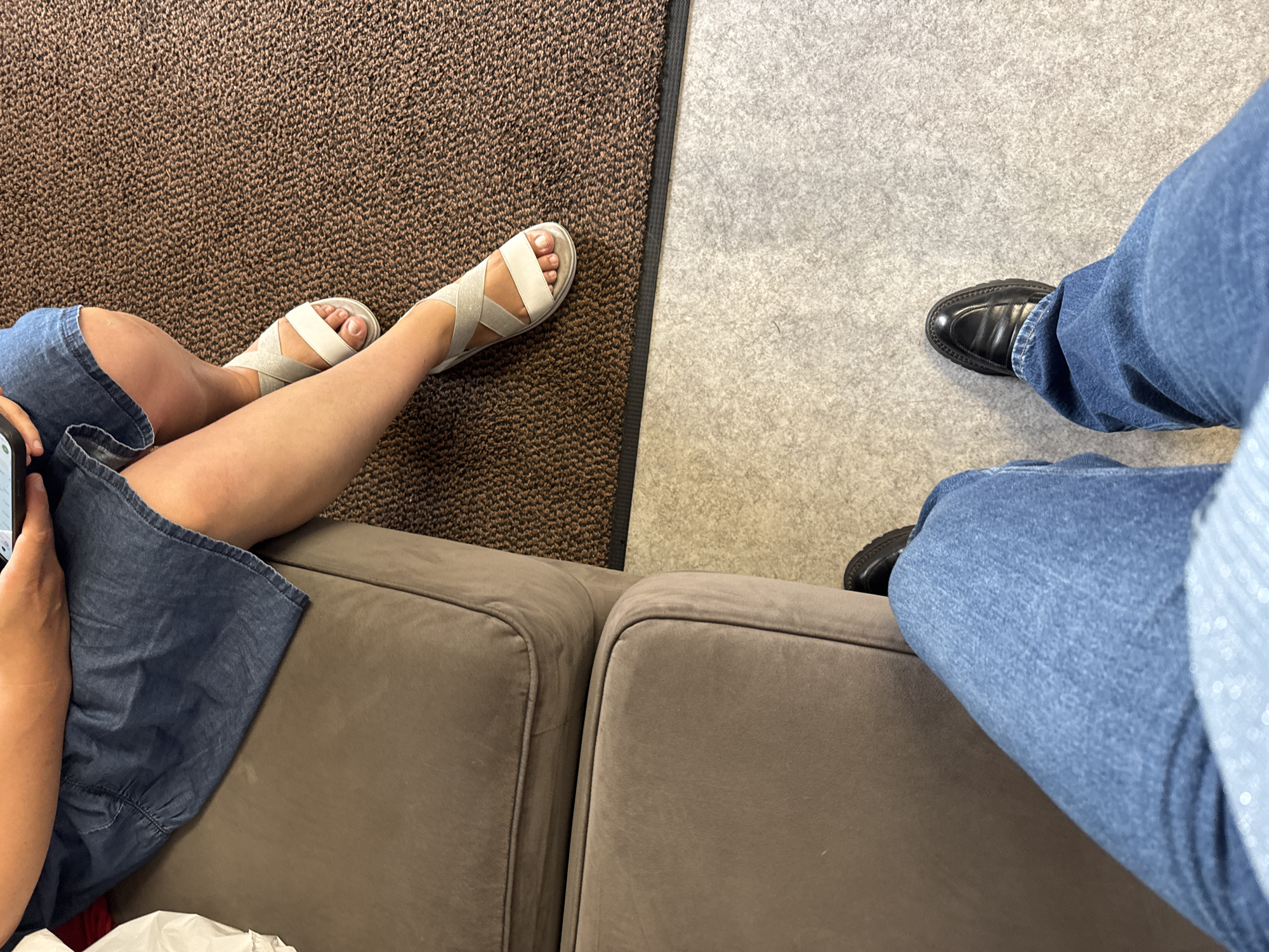 The image shows a close-up view of a person's legs and feet. One person is sitting on a brown couch, wearing a blue denim dress and white sandals. Their legs are crossed, and they are holding a phone. Next to them, another person is standing, wearing blue jeans and black loafers. The floor appears to be a combination of carpet and a lighter, speckled material. The scene is indoors, but there is no indication of the specific location within Clonmel, Ireland.