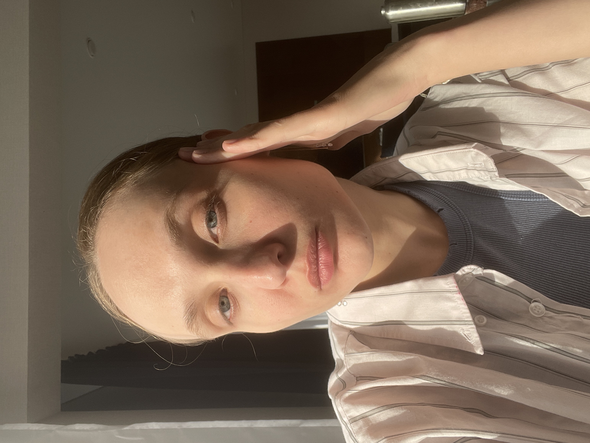 A fair-skinned person with light hair and green eyes is positioned in the foreground, with their left hand touching their forehead. The individual is wearing a gray ribbed tank top covered by a striped white and light pink button-down shirt. Light from an unidentified source casts shadows on their face and the surrounding environment. The background includes a section of a light-colored wall and a dark wooden or brown surface that could be a door or piece of furniture. There is also a metallic object, possibly a handle or rail, visible at the top of the frame. No indicators can confirm the location as Sepych, Russia.