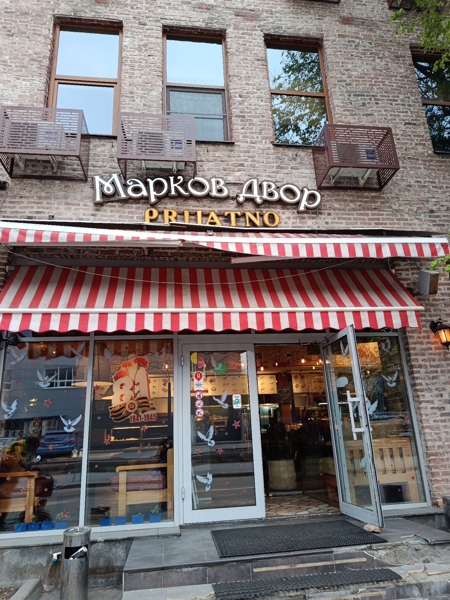 The image shows the front of a store named "Марков Двор PIIATNO" in Moscow, Russia. The building has a brick facade with four windows on the upper level, each protected by a metal cage. Below the windows is the store's sign in white letters. A red and white striped awning covers the entrance. The entrance consists of glass doors that are open, revealing the interior of the store. Decorations, including doves and stars, adorn the windows. A poster commemorating 1941-1945 is visible inside the store. A metal ashtray stands on the sidewalk in front of the store.