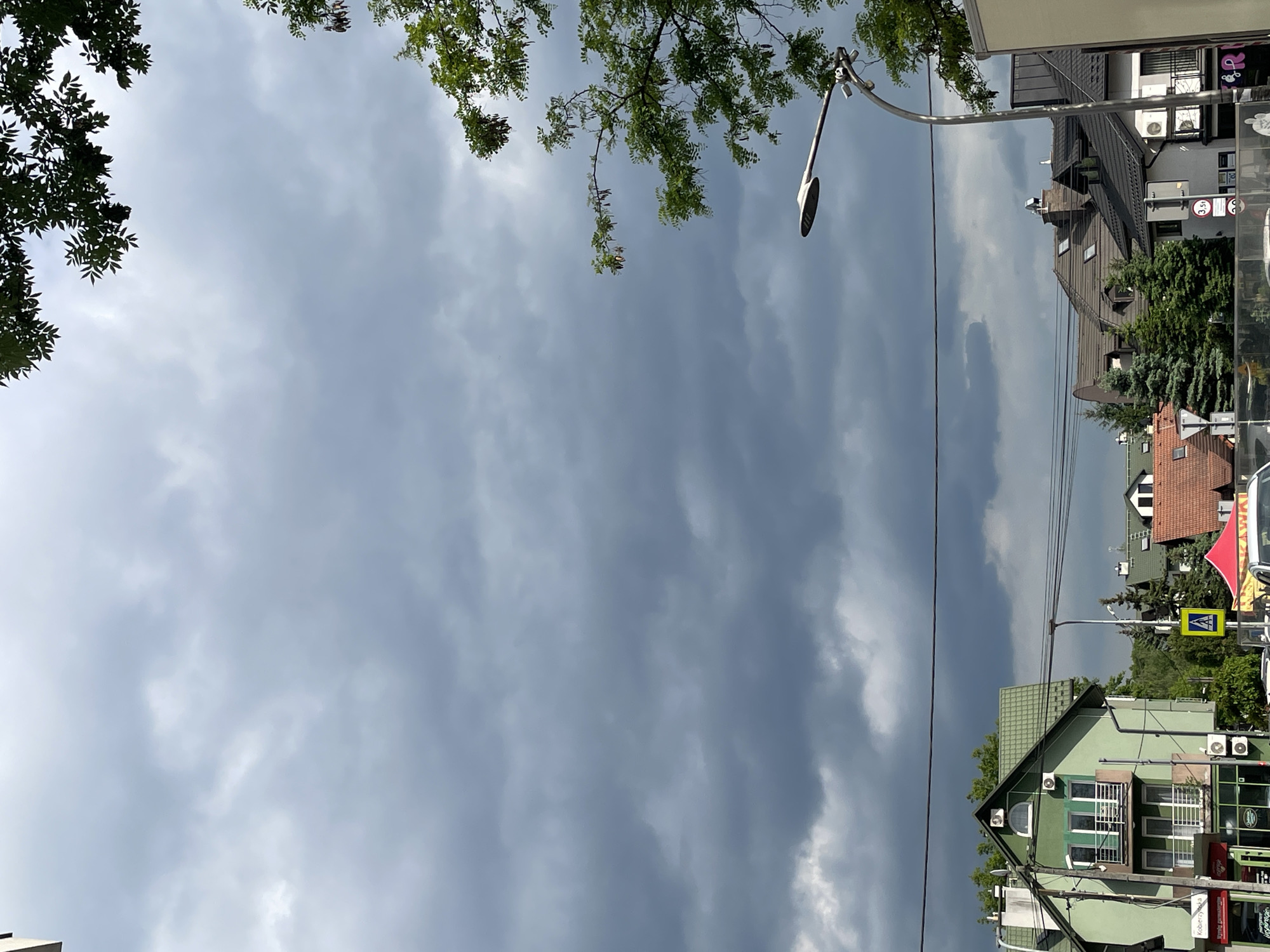 A low-angle, outdoor shot depicts a street scene in Unknown, Unknown, under an overcast sky. Dark, heavy clouds dominate the view. Trees with green foliage frame the top corners of the image.
A curved street lamp extends into the frame from the top right. Power lines stretch across the sky.
Buildings line the street. One appears green with a peaked roof and multiple windows. Air conditioning units are visible on its exterior. Other buildings with tiled roofs and windows of varying styles appear farther down the street. Sidewalk signage is visible, though not clearly legible. A pedestrian crossing sign is present. The photograph seems to have been taken from a standing position.