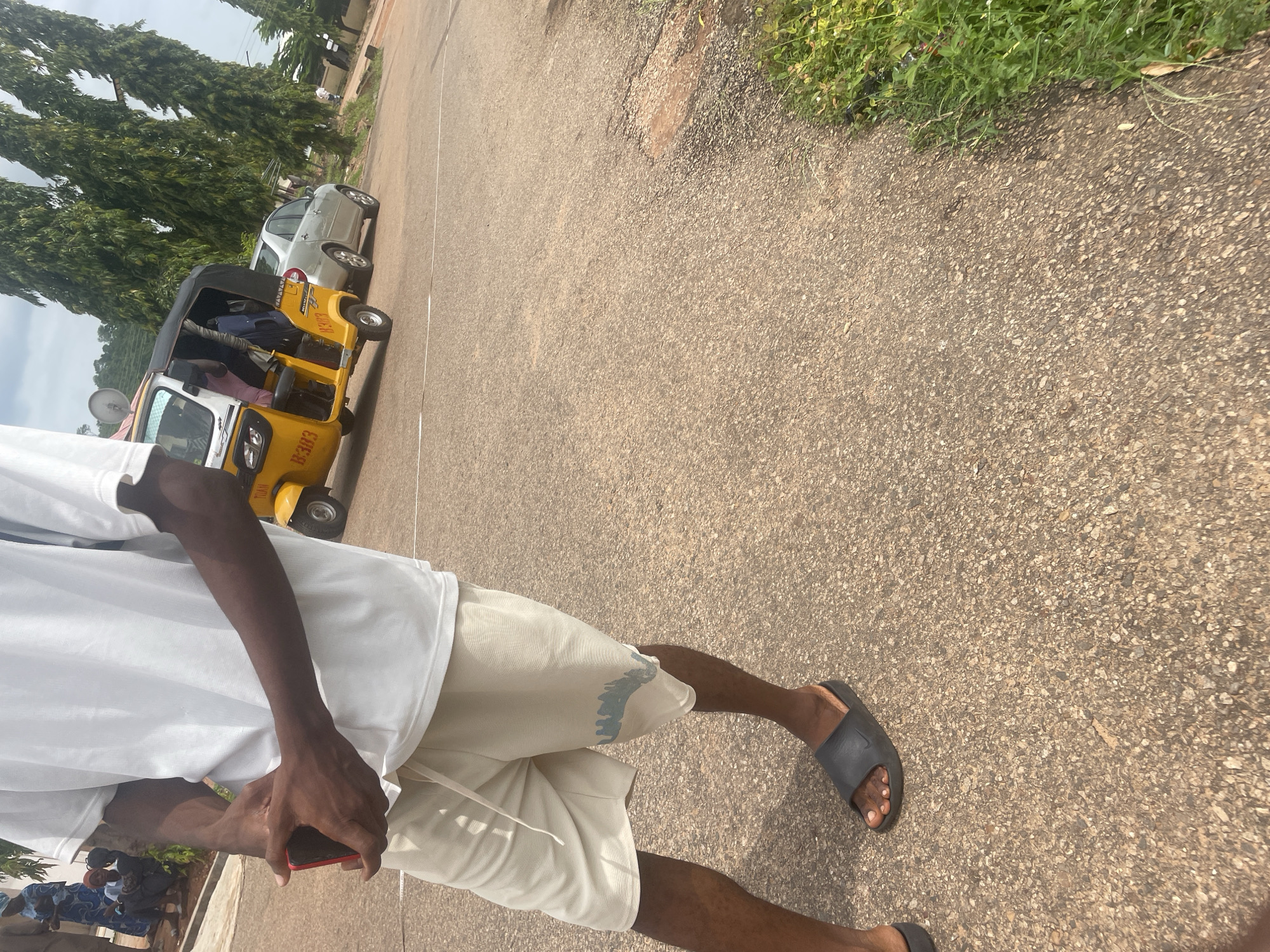The image, captured in Lokoja, Nigeria, depicts a street scene. In the foreground, a person is shown from the waist down, wearing a white t-shirt, light-colored shorts with a blue graphic design, and black slide sandals. The person is holding a red object, possibly a phone, in their left hand. Behind them, a yellow auto-rickshaw is parked on the asphalt road, with its doors open. A silver sedan is parked next to the auto-rickshaw. The background features lush green trees and a cloudy sky. In the far left of the image, several individuals are visible near a building. The environment appears to be a public road with some vegetation on the right side.