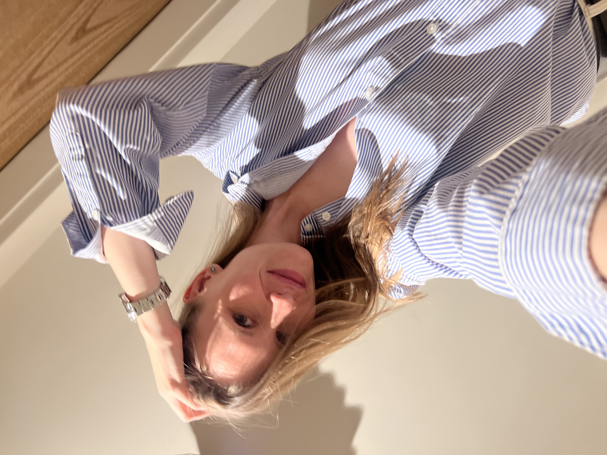 A fair-skinned woman with blonde hair is depicted from a low-angle selfie perspective. She is wearing a blue and white striped button-down shirt. A silver watch is visible on her left wrist. The background is a neutral tone, showing sections of a wall and a light-colored wooden surface.