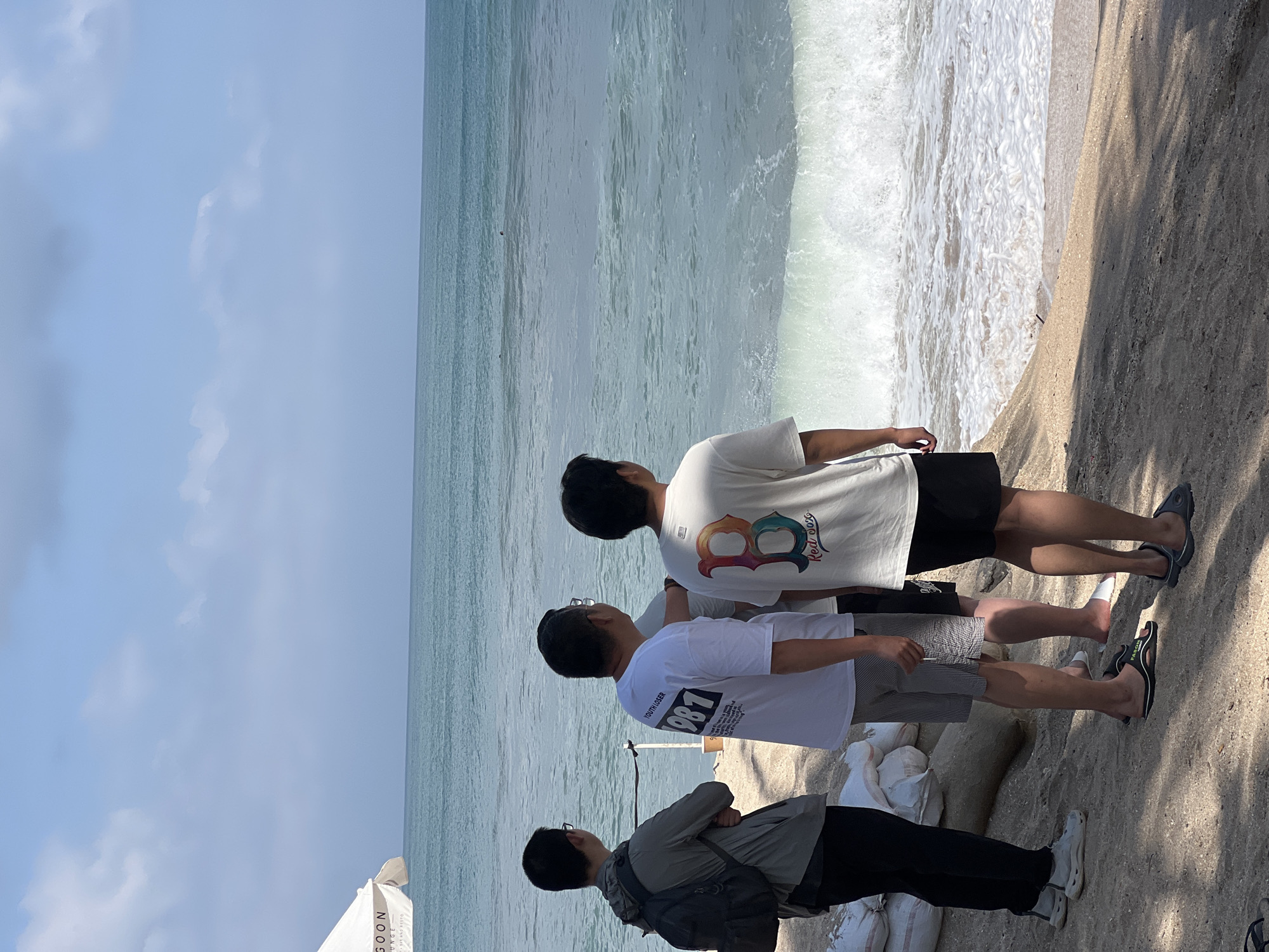 Four individuals are standing on a sandy beach, facing the ocean. Two of them are closer to the water, with waves breaking near their feet. The leftmost person is wearing a grey jacket and a black backpack. The person next to them has on a white t-shirt with "YOUTH LOSER 1987" printed on the back. The third individual is wearing a white t-shirt with a "B" logo and "Red Sox" on the back. The fourth person's back is partially visible. In the background, the ocean stretches to the horizon under a partly cloudy sky. A white umbrella, partially visible on the left, has "GOON LOUNGE - BAR AND RESTO" printed on it, suggesting a nearby establishment in Kuta, Indonesia. Several sandbags are piled up on the beach to the left of the group.