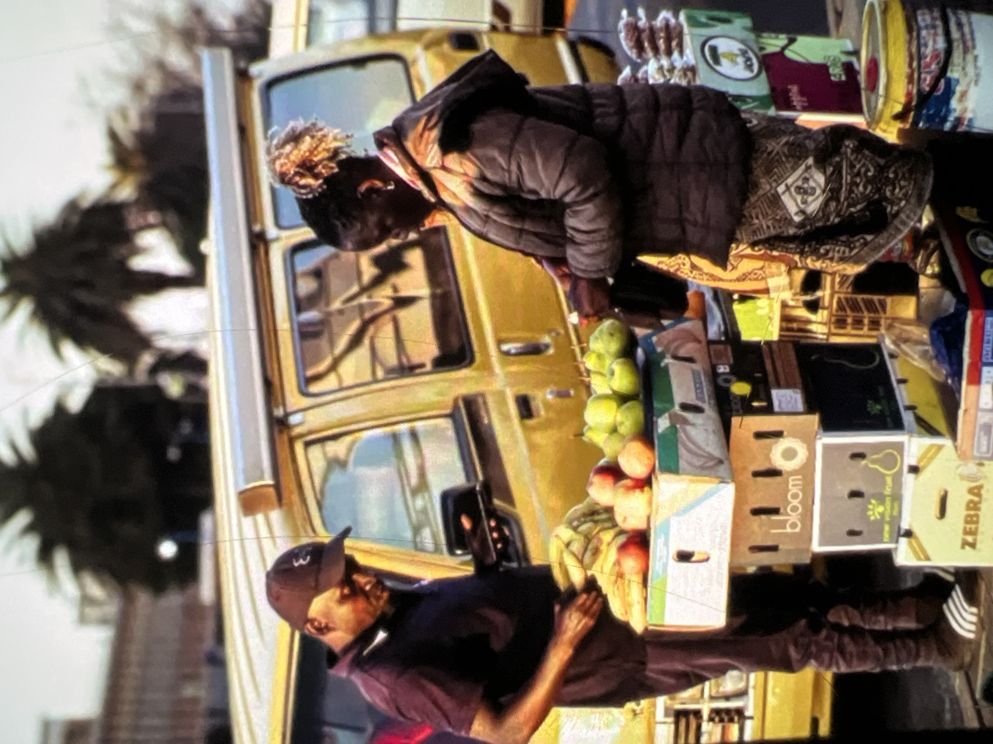 The image depicts a street scene in Lagos, Nigeria, with a focus on two individuals at a roadside fruit stall. In the foreground, a man wearing a dark t-shirt, dark trousers, and a baseball cap is looking down at a display of fruit. He is standing next to stacks of cardboard boxes filled with produce, including apples and pears. Behind him, a yellow van is partially visible, suggesting a busy urban environment. To the right, a woman in a dark quilted jacket and a patterned skirt is interacting with the fruit stall. She is holding a small bag and appears to be making a purchase. The stall itself is laden with various fruits, and in the background, other market stalls and palm trees are discernible, typical of the Lagos landscape. The lighting suggests it is daytime, with a bright, overcast sky.