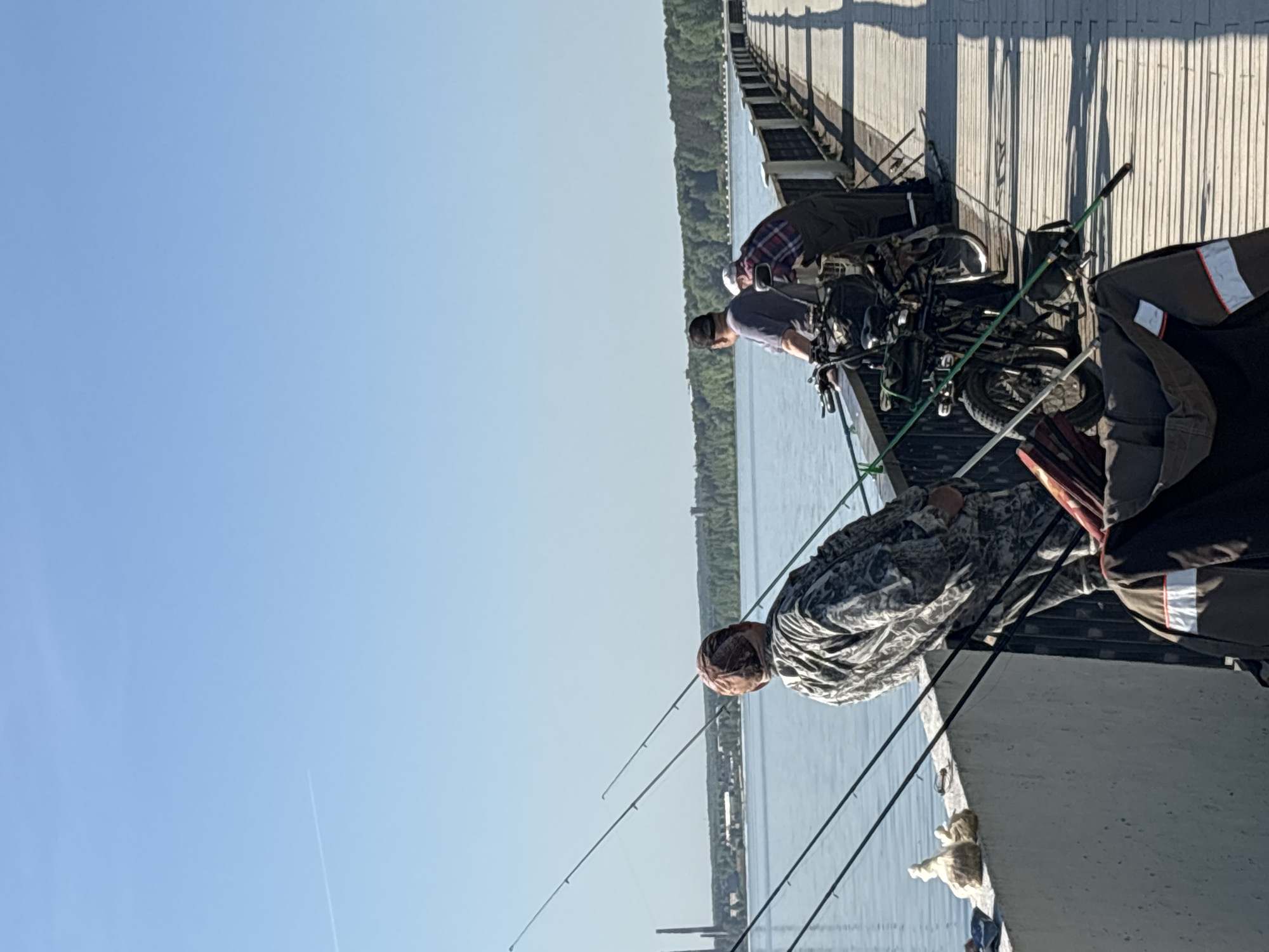 The image depicts three men fishing from a concrete pier or bridge. The men are positioned in a row, with fishing rods extending out over the water. A motorized bicycle is parked behind the men. The water is calm and stretches to the horizon, where a line of trees is visible. The sky is clear and blue. The location is likely a pier in the city of Chaikovsky, Russia.