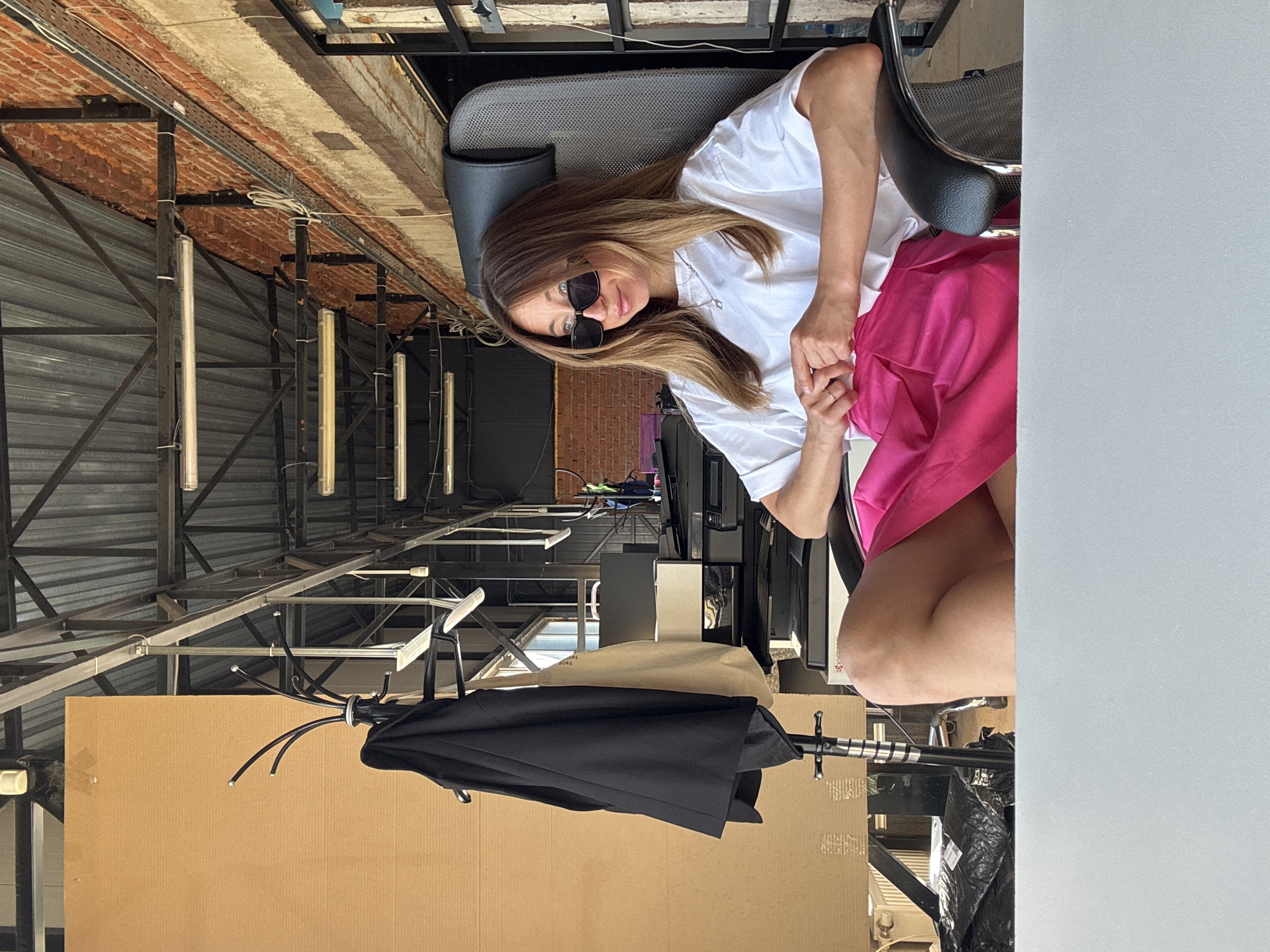 A woman wearing sunglasses, a white t-shirt, and a pink skirt sits in an office chair in what appears to be an industrial or commercial space in Chaikovsky, Russia. The space has exposed metal framework, corrugated metal walls, exposed light fixtures, and a brick wall. Near her is a cardboard box, an umbrella, a beige bag, and office equipment. The scene suggests a temporary or unconventional workspace.