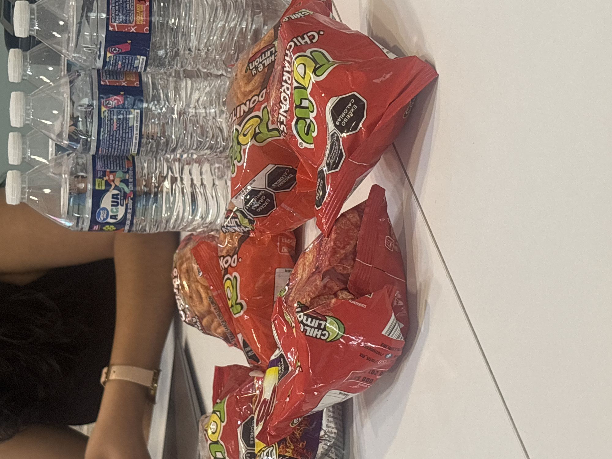 The image shows a partial view of a person's left arm wearing a watch, alongside a stack of water bottles and several bags of Donitas Chile y Limón-flavored wheat snacks. The water bottles appear to be "Great Value" brand. The snack bags have nutritional information printed on them, including calorie and fat content. The text on the bags suggests the product is made in Mexico. The image also indicates the snacks are available for purchase at a "COSTCO" location, based on the text near the bottom of one of the bags. No other specific details about the location in Buenavista, Mexico, are visible in the image.