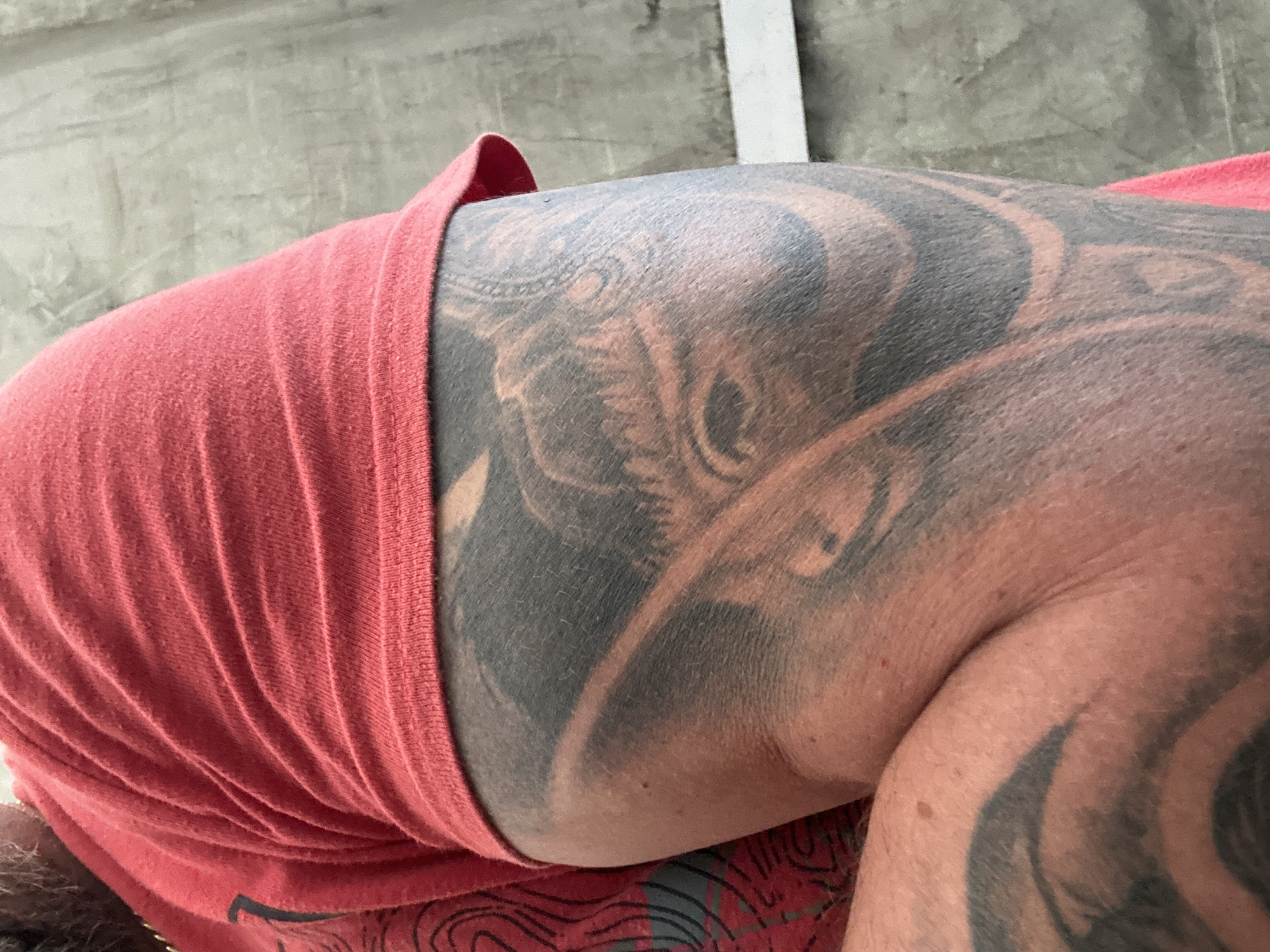 The image is a close-up of a person's arm, which is covered in a large tattoo. The person is wearing a red t-shirt. The background appears to be a textured wall. There is no information within the image that indicates the location in the city of Dornbirn, Austria.