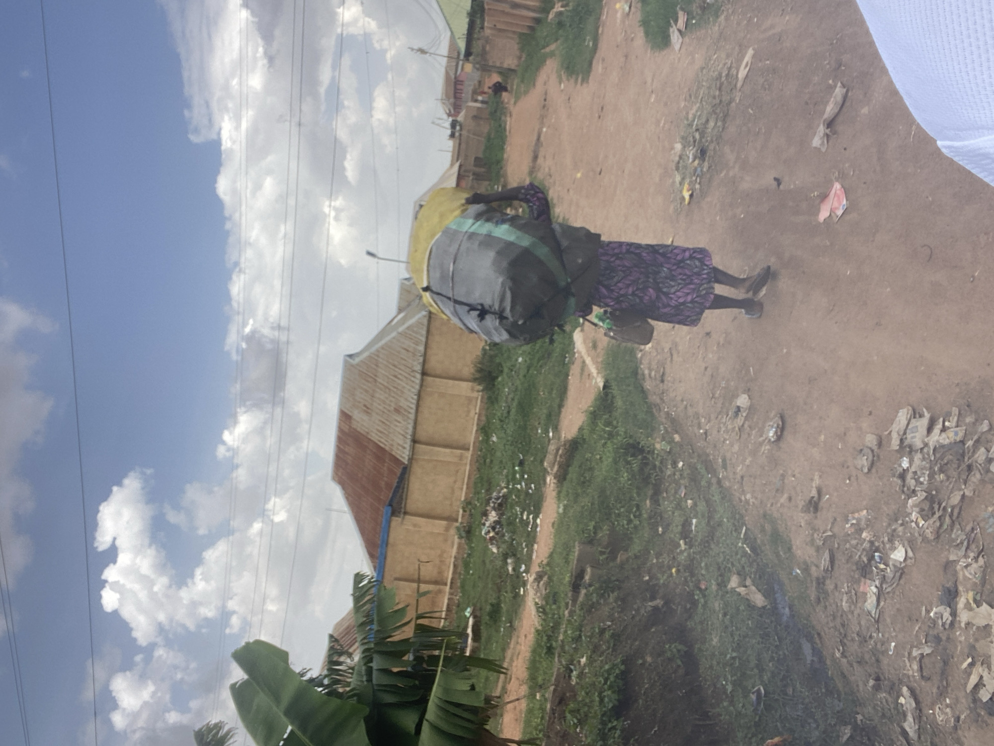 An adult is carrying a large bundle atop her head along a dirt path in Bukuru, Nigeria. The bundle consists of what appears to be two large bags secured together. The ground shows signs of scattered debris and vegetation. Buildings are visible in the background. A blue sky with white clouds is overhead. Banana trees are in the foreground. Power lines are visible in the upper left corner.