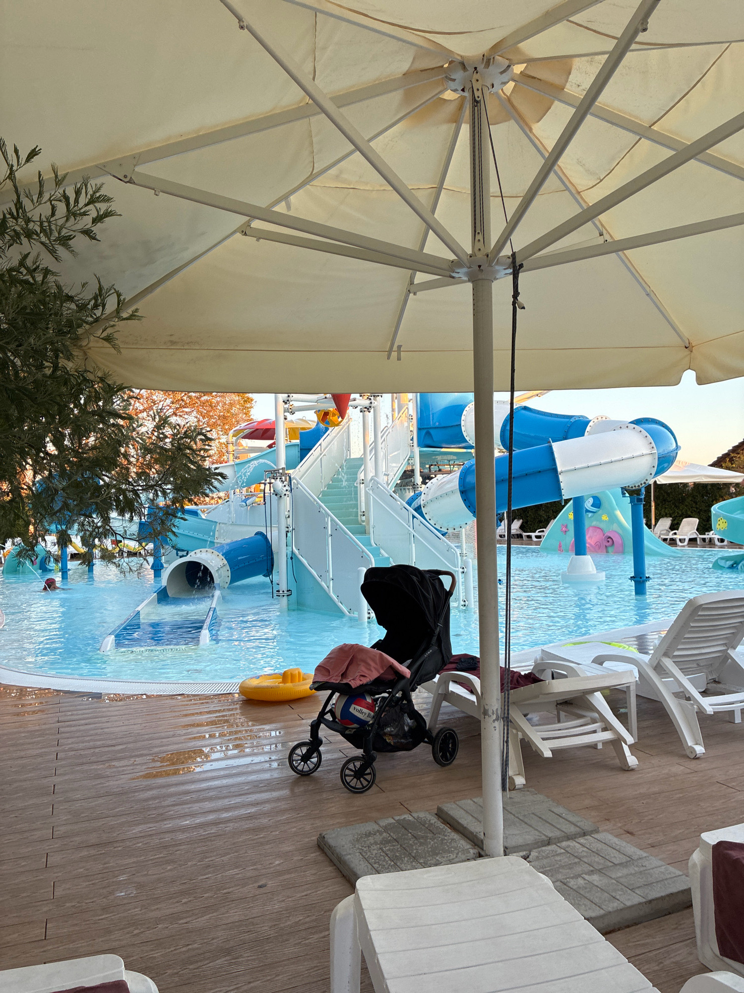 The image captures a water park with blue and white water slides. A person is visible in the pool on the left side of the image. In the foreground, a black baby stroller with a pink blanket and a volleyball is parked on a wooden deck. White lounge chairs are also present on the deck. A large umbrella shades a portion of the deck. The water park appears to be located in the city of Sochi, Russia.