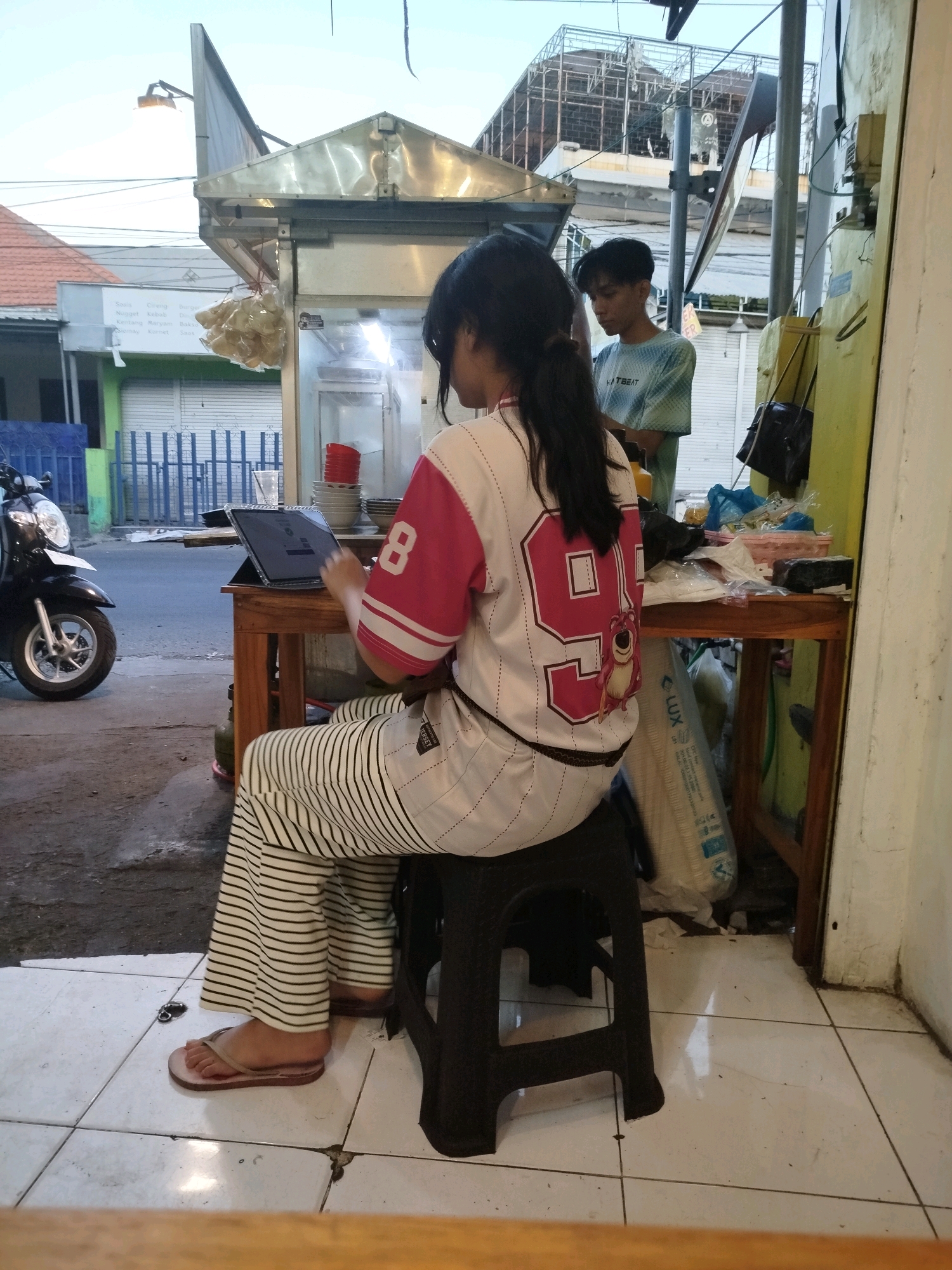 The image depicts a street food stall in Surabaya, Indonesia, with two individuals. In the foreground, a young woman with long dark hair pulled into a ponytail is seated on a black plastic stool, facing away from the camera. She is wearing a white and pink jersey-style top with the number "99" in a large pink square, and a cartoon character on the back. Her pants are striped black and white. She is barefoot, wearing light-colored flip-flops. A tablet computer is positioned on a wooden table in front of her, and her right hand appears to be interacting with it.
Behind her, slightly to the left and in the mid-ground, a young man with short dark hair is standing. He is wearing a light green and blue tie-dye t-shirt with text that appears to read "HYPEBEAST". He is looking down.
The stall itself is a simple structure with a metal roof. Bags of goods are visible behind the table, including a large white bag with blue writing that says "LUX". Various food items, possibly snacks, are hanging in a plastic bag from the roof of the stall. A black scooter is parked to the left of the scene. The ground is paved with white tiles. In the background, buildings and a partially constructed structure are visible, suggesting an urban environment. The lighting indicates it is either early morning or late afternoon.