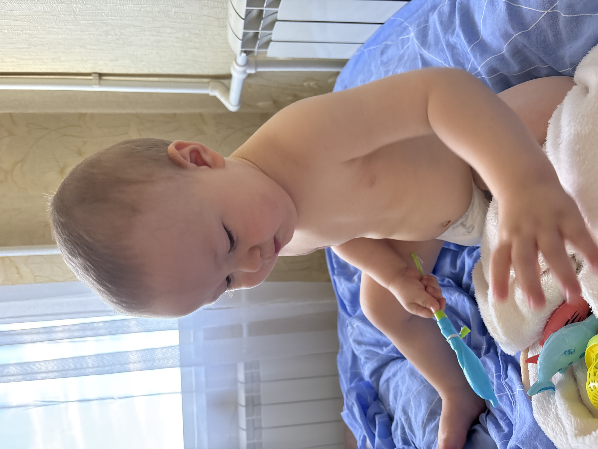 A baby is sitting on a blue blanket on a bed, playing with toys. The baby is wearing a diaper. There are white curtains and a radiator in the background. The scene appears to be indoors, likely in a bedroom. There are no indications of the specific location within Naberezhnye Chelny, Russia.