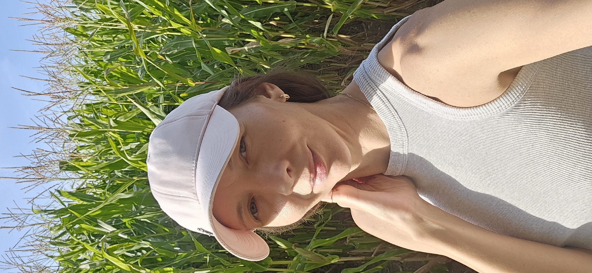 The image features a woman lying down in a cornfield. She is wearing a light-colored baseball cap and a ribbed tank top. Her right arm is bent, with her hand resting near her neck and chin, and a ring is visible on one of her fingers. She has a gold hoop earring in her left ear and a delicate gold necklace around her neck. The woman's face is partially obscured by the cap, but her eyes are open, and she appears to be smiling softly.
The setting is a sunlit cornfield, with tall green corn stalks filling the background and extending towards the top of the frame. The sky is a clear blue. The lighting suggests it is daytime, likely late morning or early afternoon, given the angle of the sun and the shadows. The image is oriented vertically, with the woman positioned from the upper left to the lower right of the frame. The overall mood is relaxed and serene, with the woman enjoying the natural environment. No visible text is present in the image.